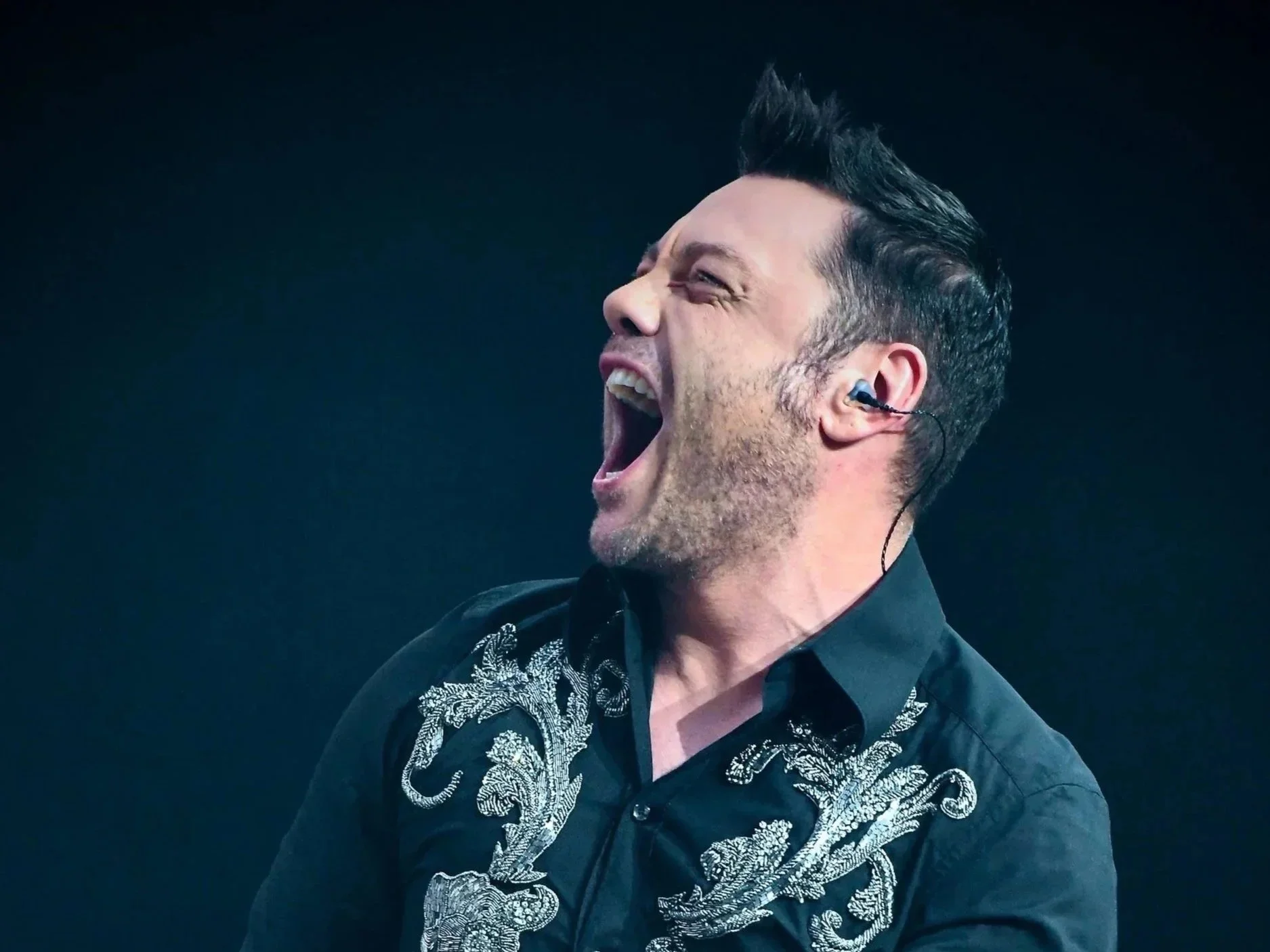 Un uomo canta con entusiasmo, indossando un auricolare e una camicia nera decorata con motivi di dragone argentati.