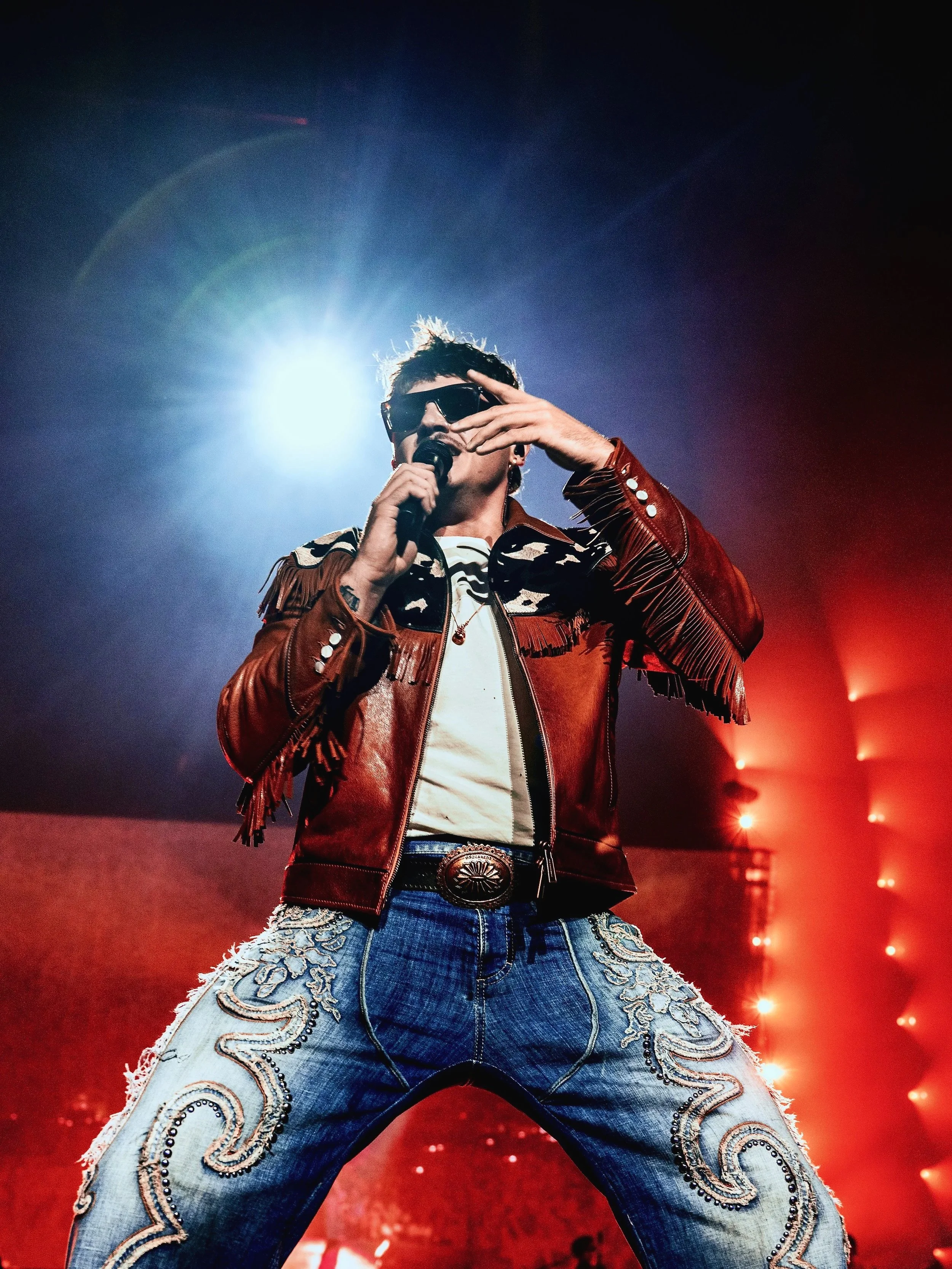 Un artista vestito con giacca di pelle rossa, jeans con ricami e occhiali da sole, tiene un microfono sul palco con luci rosse e blu sullo sfondo.