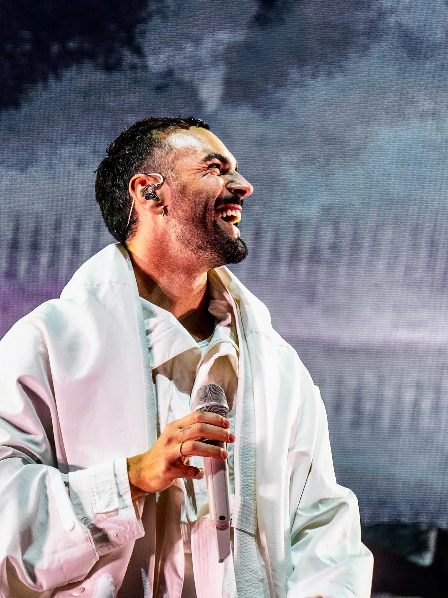 Un uomo sorridente che tiene un microfono, indossa un abbigliamento bianco, con brevi capelli scuri e barba, e ha un dispositivo auricolare.