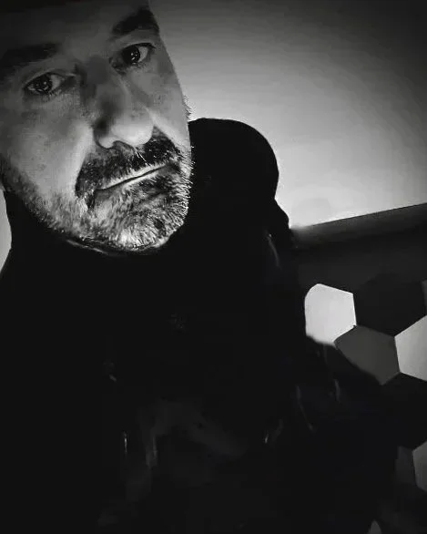 Un uomo con barba e baffi, in un ambiente in bianco e nero, guarda verso la fotocamera.