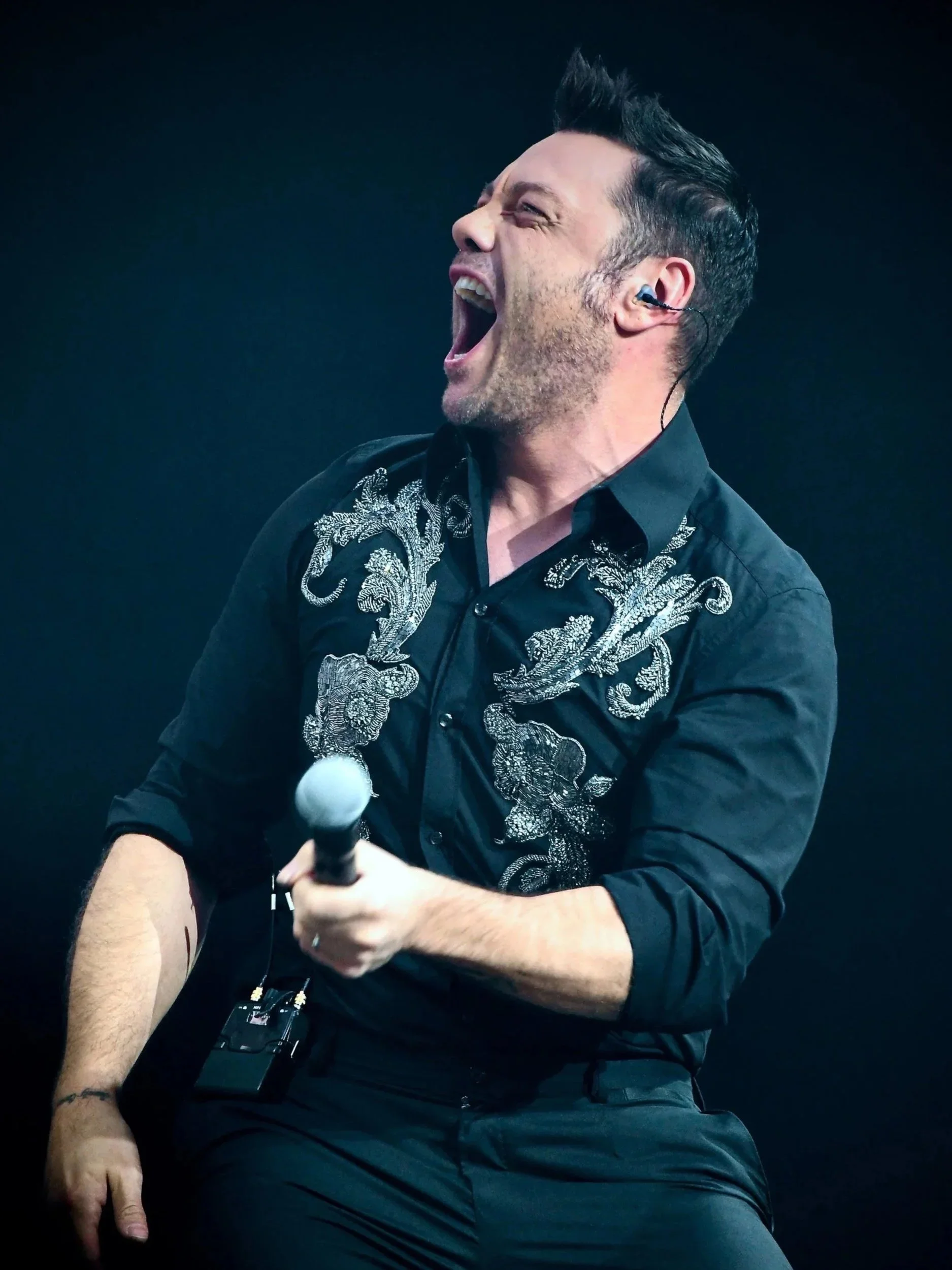 Un uomo che canta con entusiasmo durante una performance dal vivo, con in ear monitor e tenendo un microfono, indossa una camicia nera con dettagli ricamati.