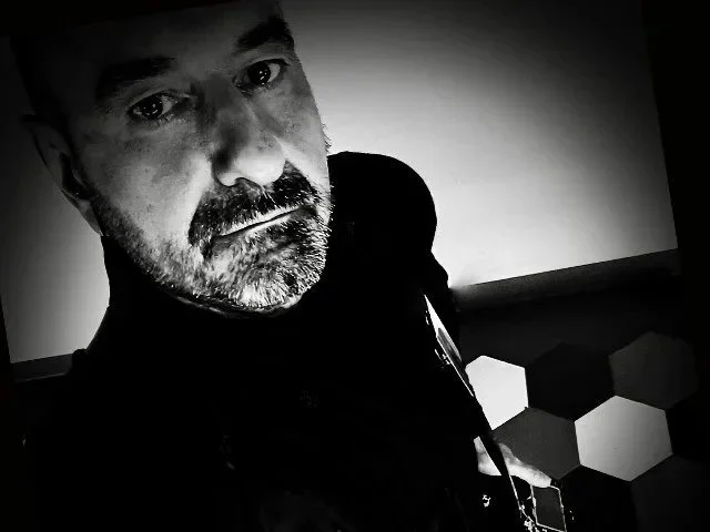 Un uomo con barba e baffi, sguardo serio, fotografia in bianco e nero, sfondo con un muro bianco e motivi a nido d'ape.