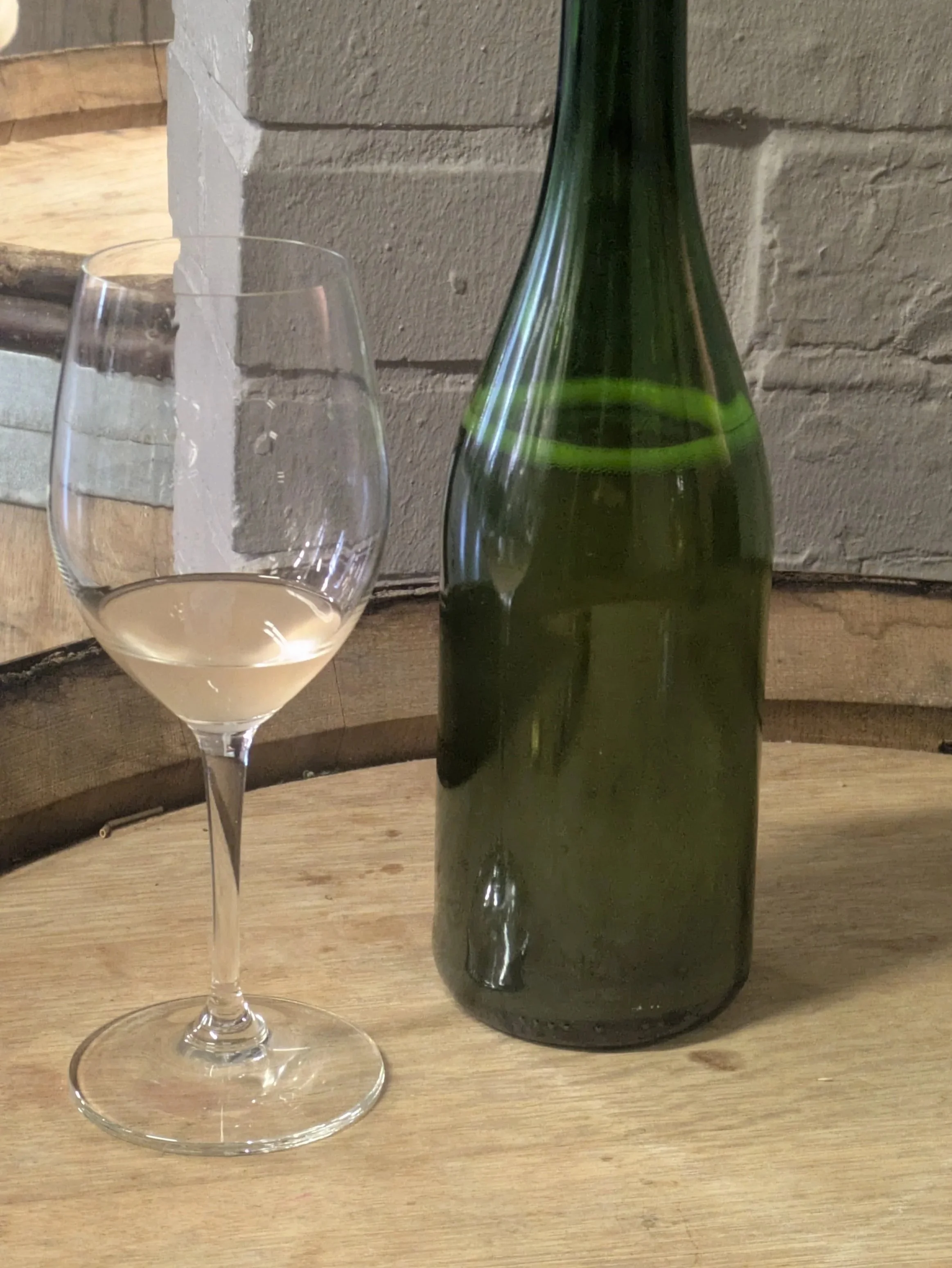 An open wine bottle next to a wine glass on an upturned barrel in front of a grey brick wall.
