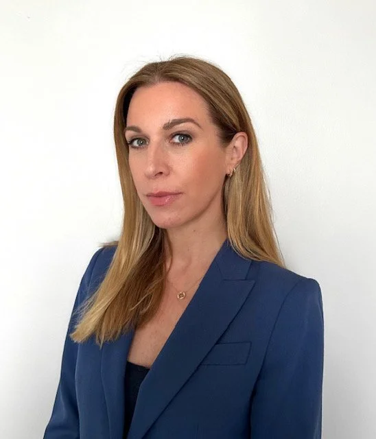 A Geography tutor with long, blonde hair, wearing a blue blazer and a black top, standing against a plain white background.