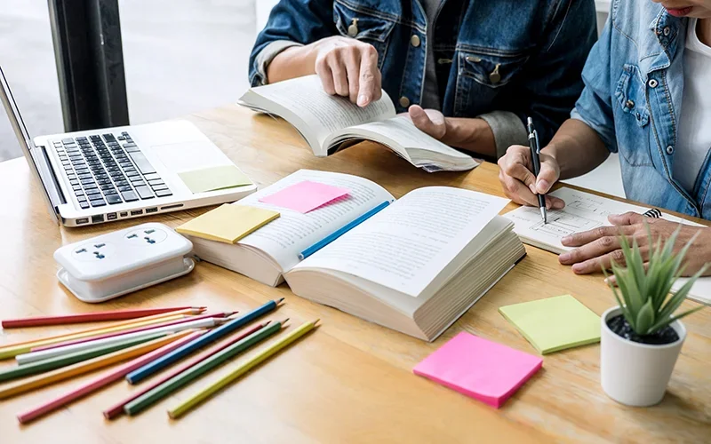 Two people studying at a desk with books, a laptop, colored pencils, sticky notes, a plant, and paper notes.