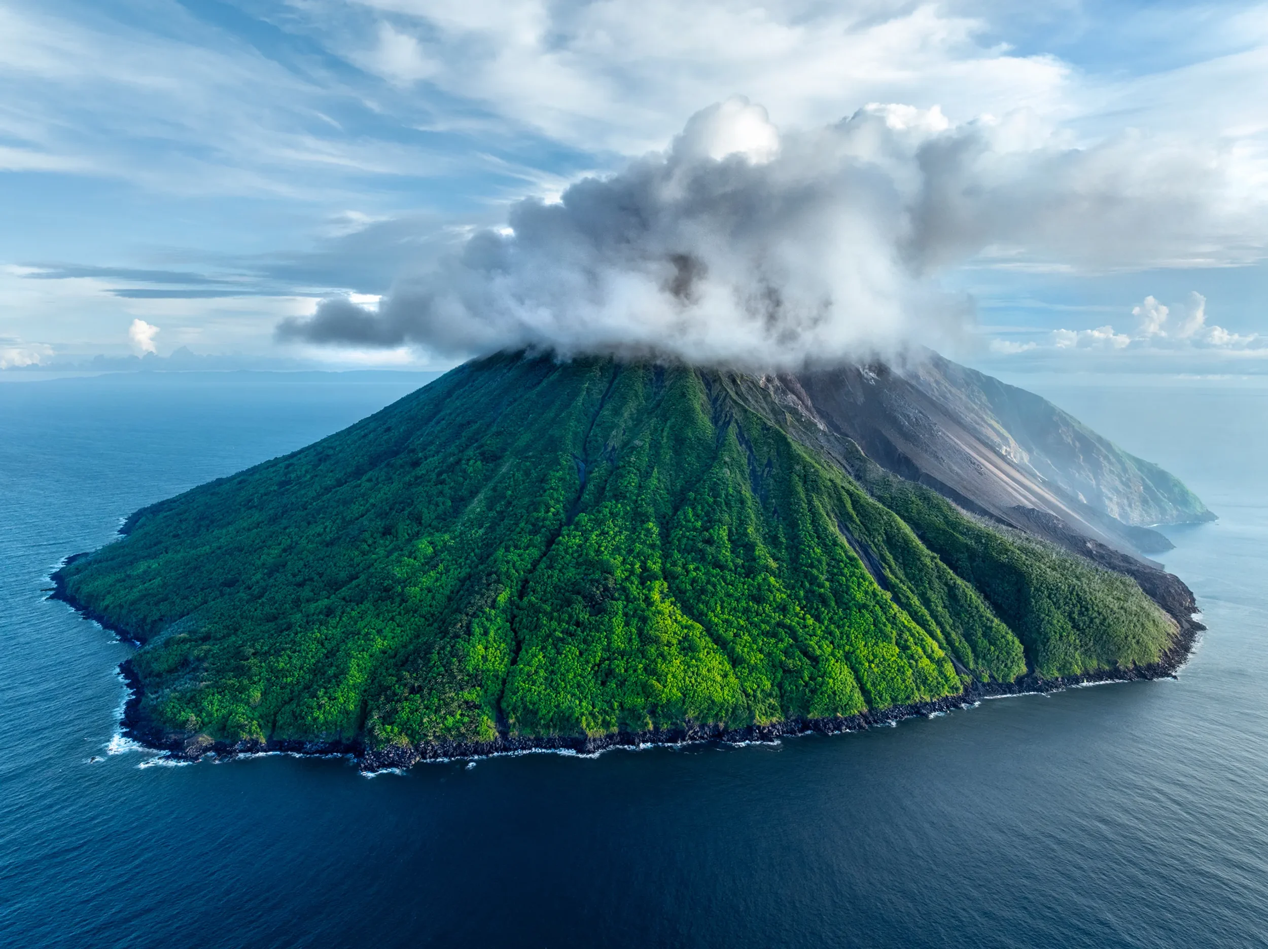 A large island in the ocean with lush green foliage on the slopes and a volcanic mountain at its center, emitting a cloud of smoke or steam into the sky.