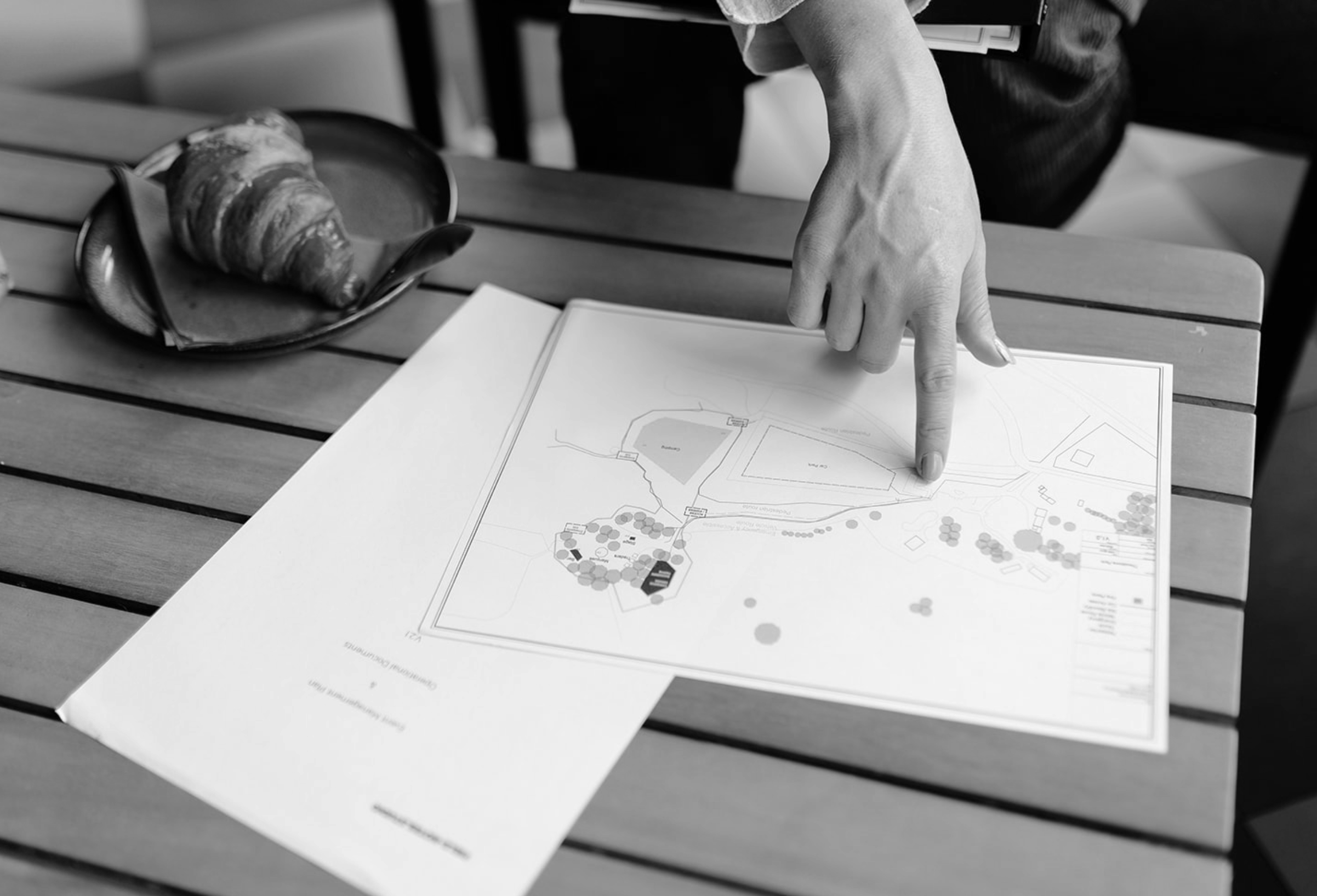 An operation manager's hand pointing to a festival site plan on a table, with a croissant on a plate nearby.