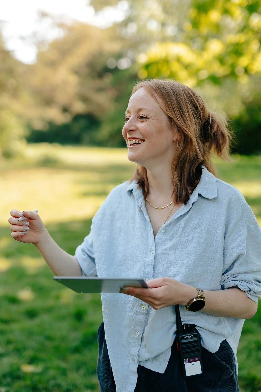A woman outdoors in a park, smiling and holding a tablet, wearing a light blue shirt, with a walkie-talkie clipped to her waistband.