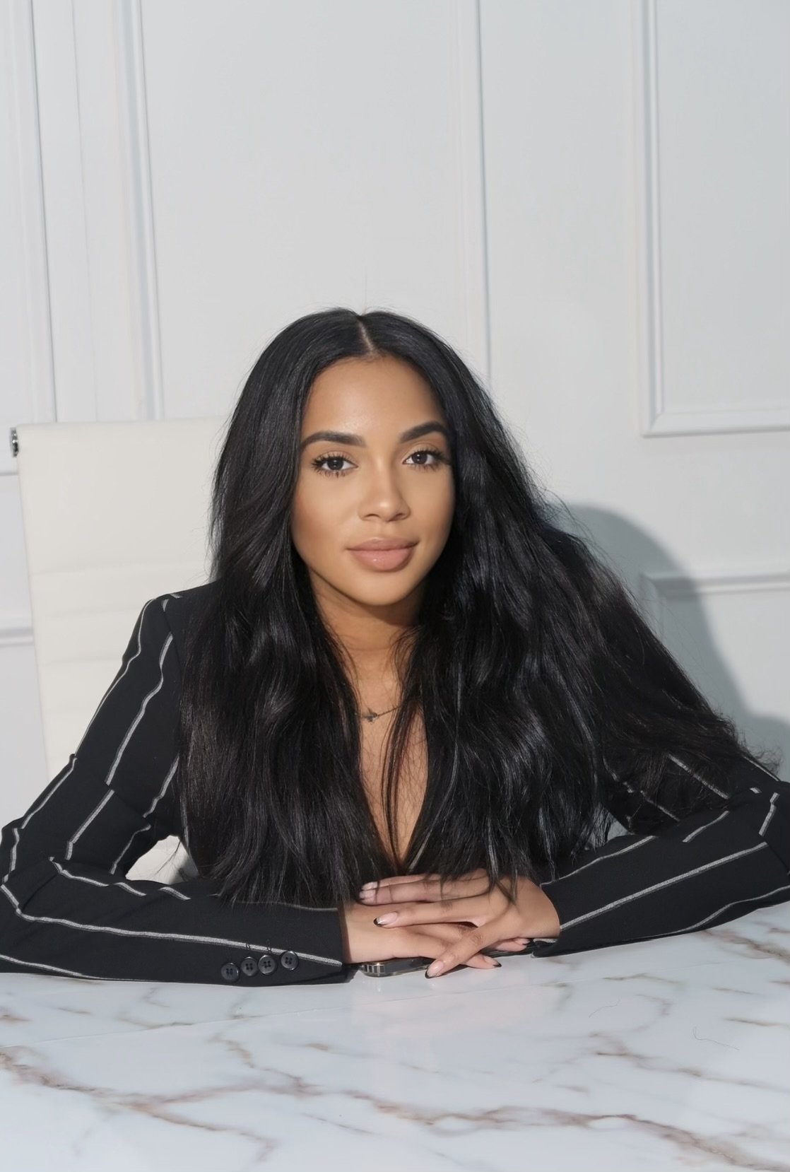 A woman with long black hair, wearing a black blazer with white stripes, sitting at a marble table in a white room.
