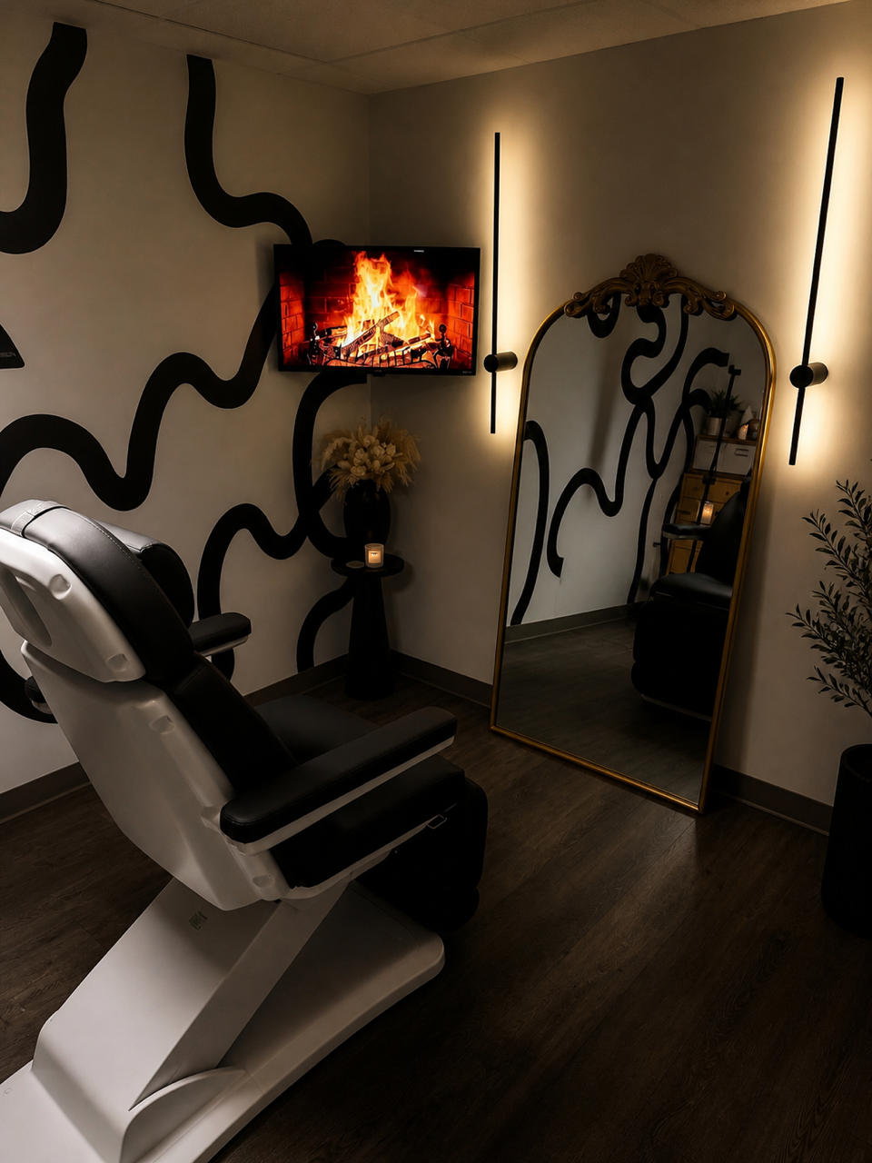 A room with a black and white wall design, a white reclining chair, a small table with a candle and flowers, a tall mirror, a wall-mounted TV displaying a fireplace, and soft ambient lighting.