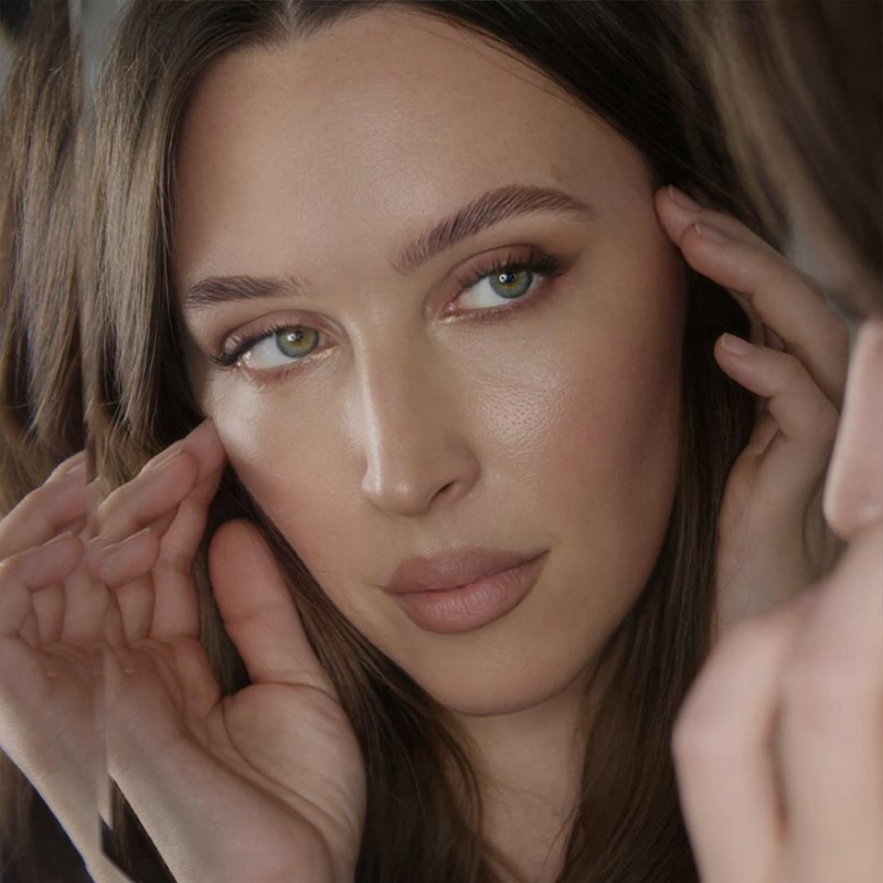 Close-up of a woman with brown hair and green eyes, touching her face gently.
