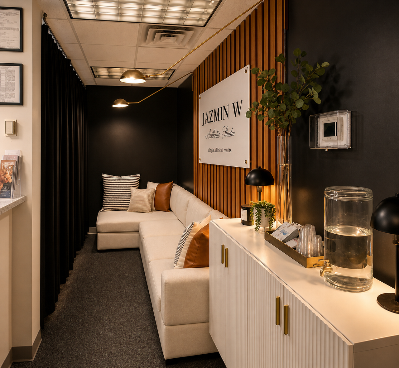 Waiting area with white sectional sofa, striped and brown pillows, black curtains, pendant lights, potted plant, water dispenser, and a sign that reads Jazmin W Aesthetic Studio.