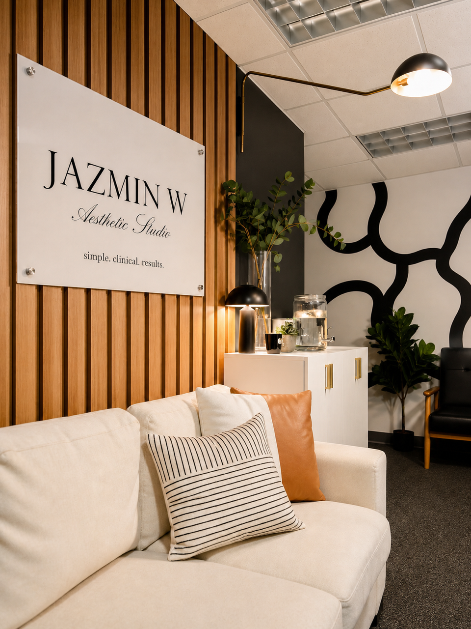 Waiting area with a cream sofa, decorative pillows, wooden wall with sign reading 'JAZMIN W Aesthetic Studio,' black wall art, plants, and modern decor.