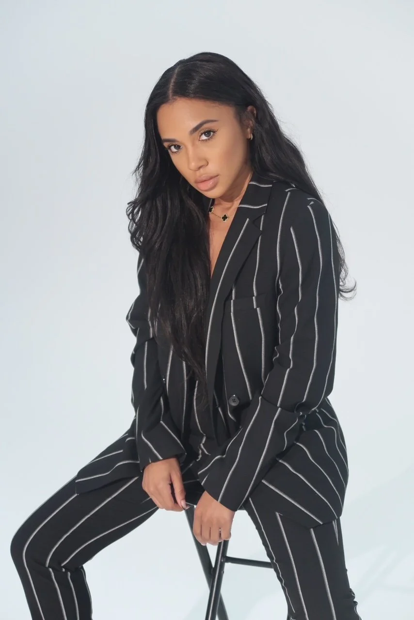 A woman with long black hair wearing a black pinstripe suit seated on a stool against a plain light background.