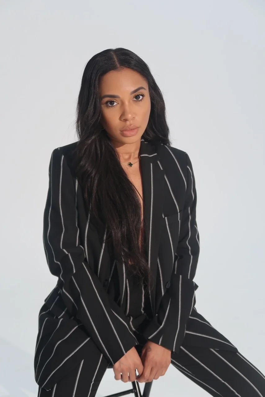 A young woman in a black pinstripe suit sitting on a stool against a plain white background.
