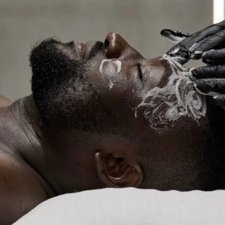 A person lying down with eyes closed while a second person applies a face mask with white cream on their face, wearing black gloves.