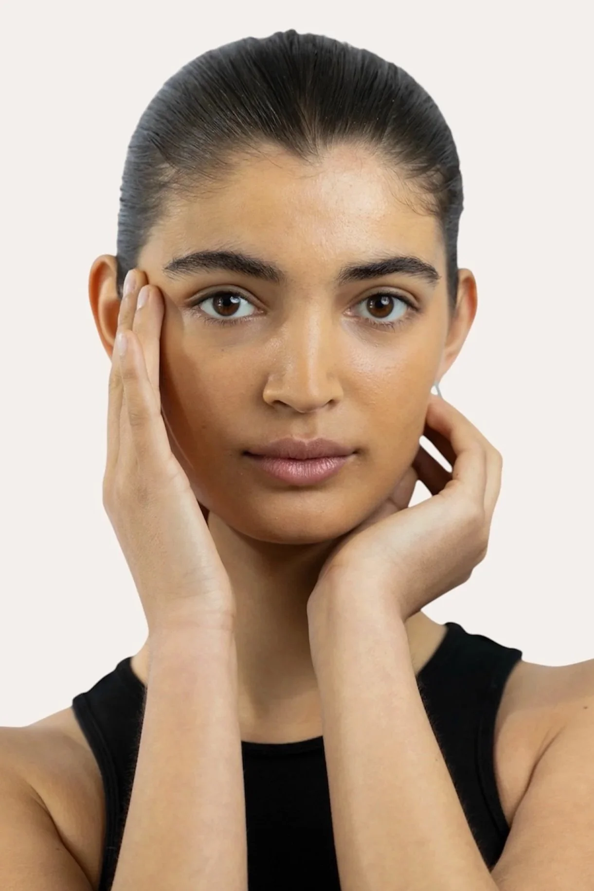 A young woman with dark hair styled in a slicked-back bun, wearing a black sleeveless top, holds her face with both hands, looking directly into the camera against a plain light background.