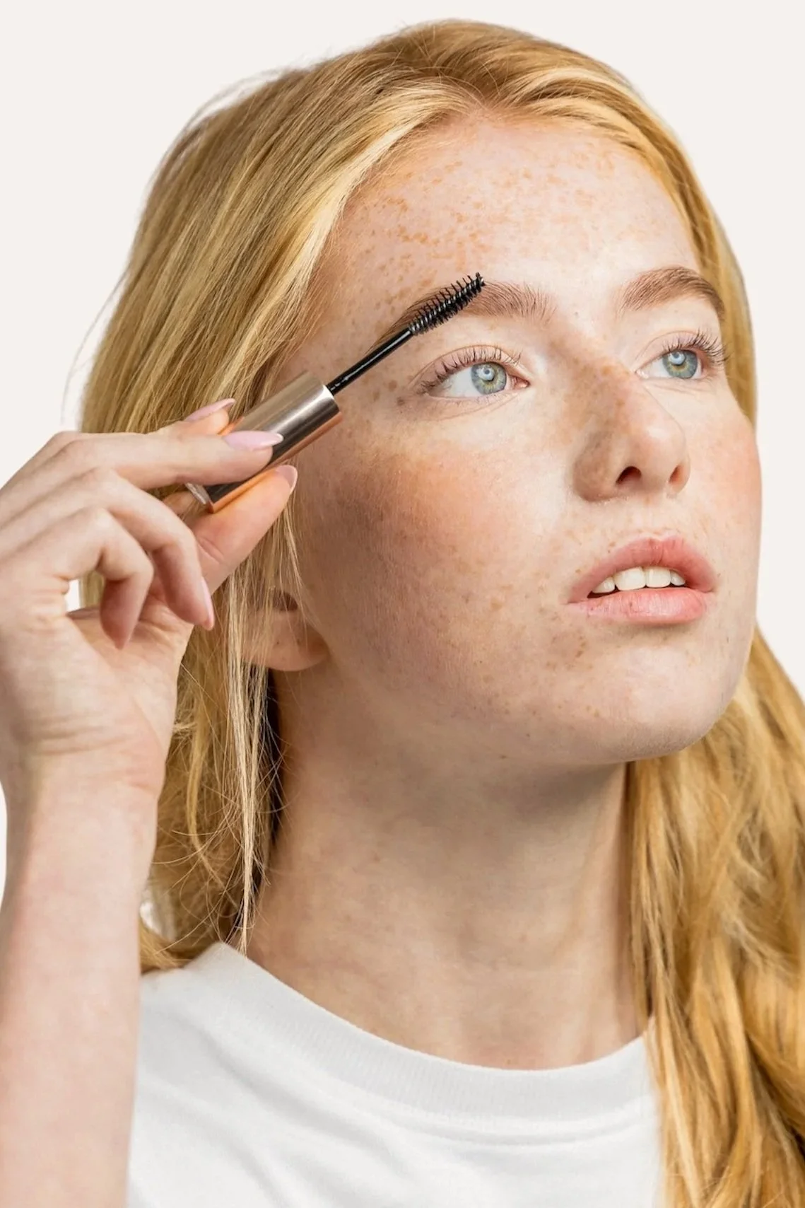 A woman with light skin and red hair with freckles is holding a mascara wand near her eyebrow, applying makeup.