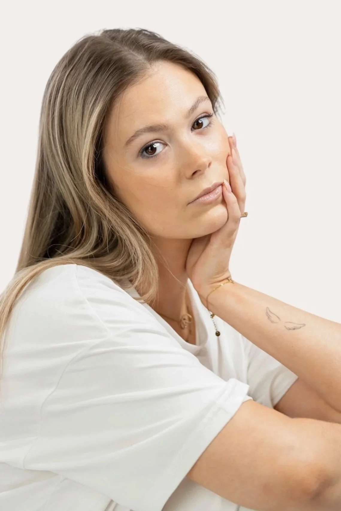 A woman with long, light brown hair, wearing a white shirt, is resting her head on her hand against a plain white background.