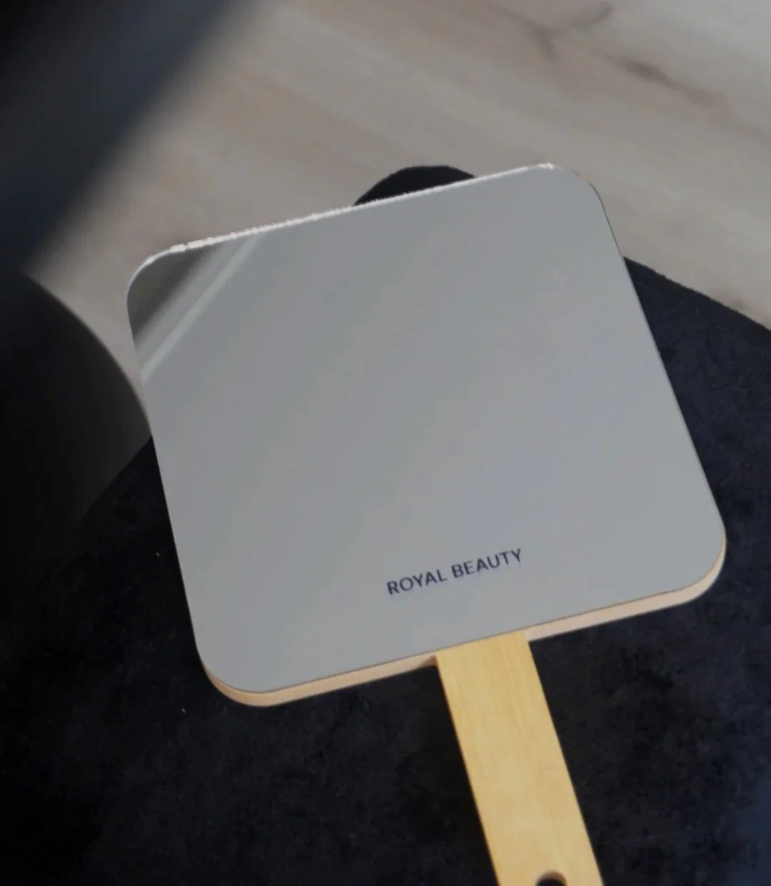 A handheld mirror with a white frame and a wooden handle, labeled 'ROYAL BEAUTY' on the back, resting on someone's lap.