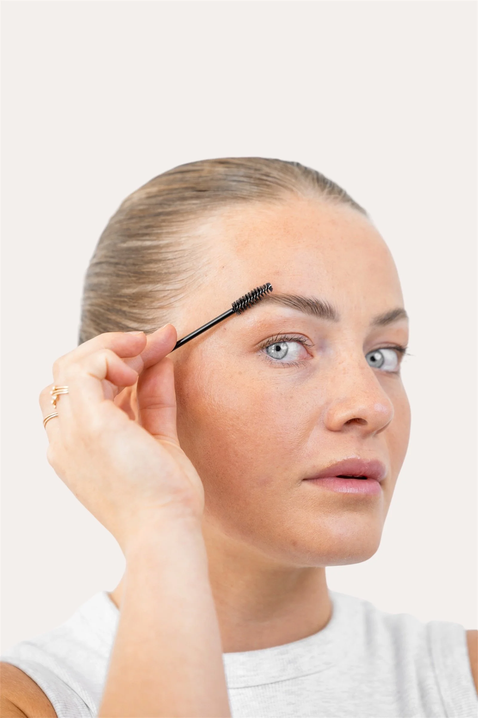A woman with blonde hair and light skin applying mascara to her eyelashes with a black mascara wand.