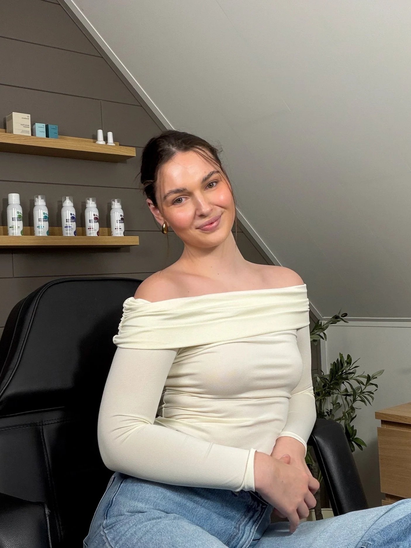 A woman with dark hair, wearing a white off-the-shoulder top and blue jeans, sitting on a black chair in a room with wooden shelves and green plants.