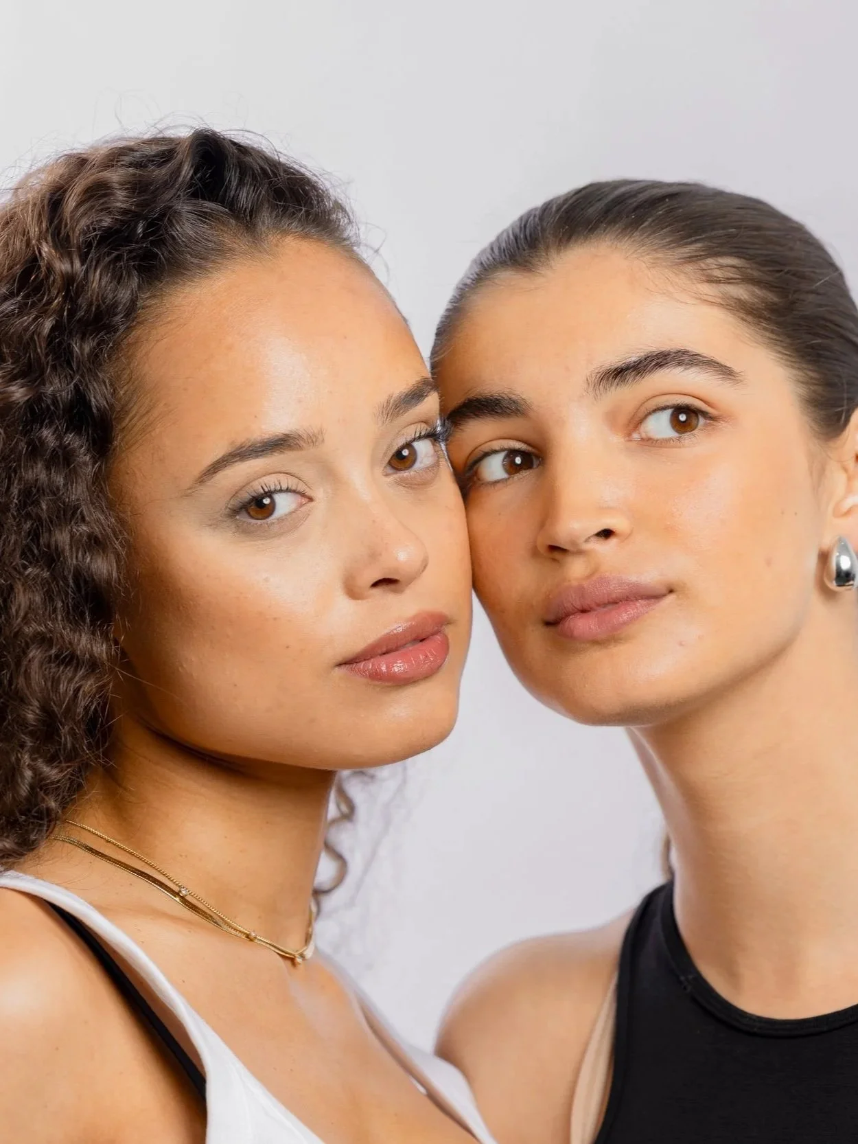 Two young women with brown eyes and natural makeup, posing closely together against a plain light background.