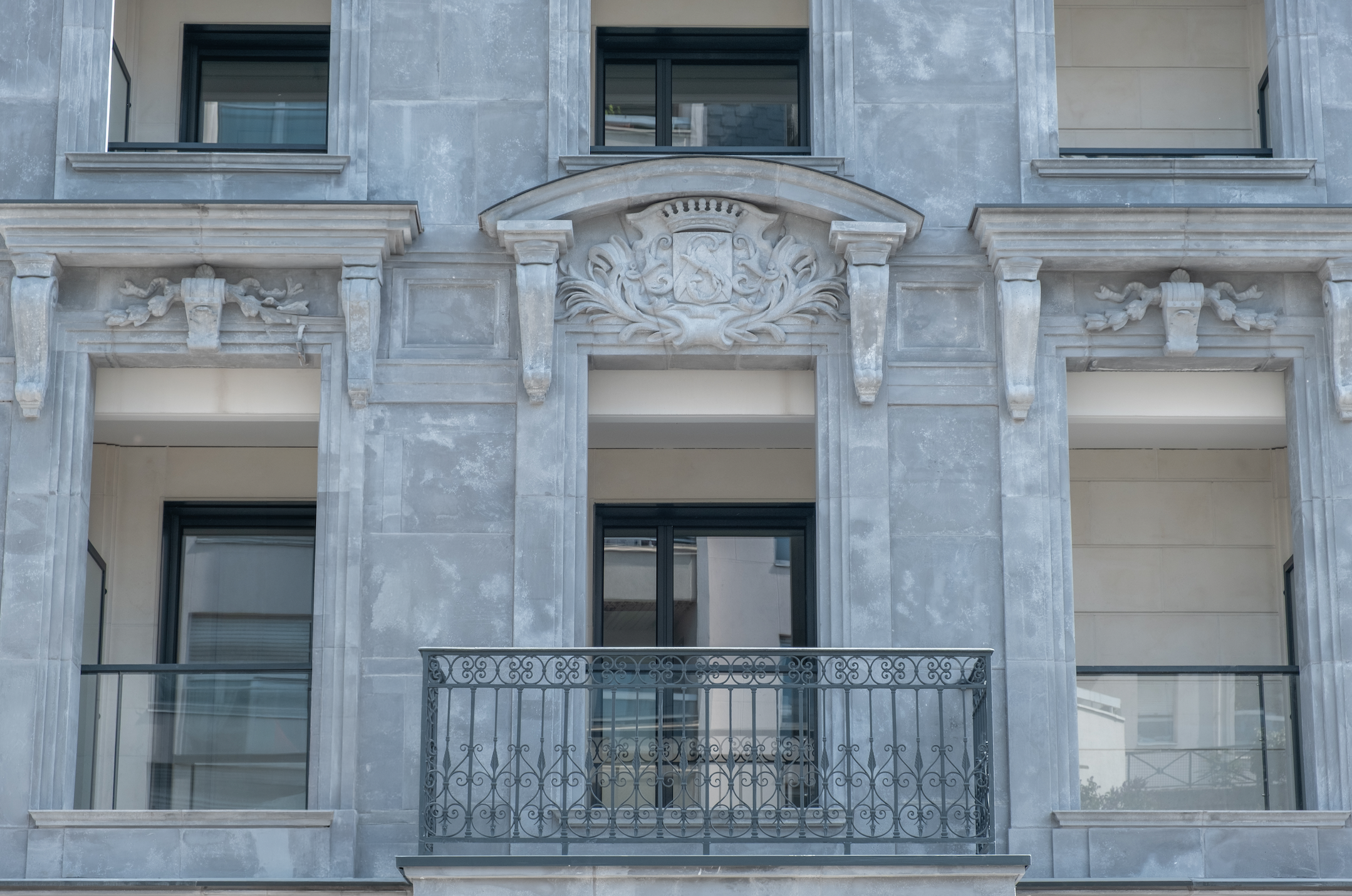 Facade of a building with gray stone exterior, featuring ornate architectural details including a crest with lions and floral elements above the balcony, and large windows.