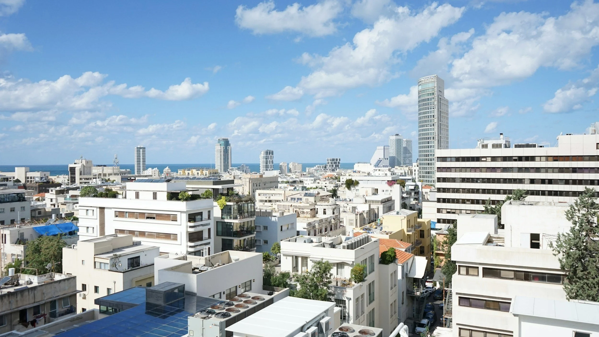 Cityscape with modern high-rise buildings, some with glass facades, and a clear blue sky with scattered clouds, overlooking the coastline in the distance.