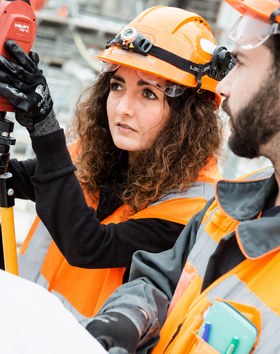 Two construction workers, a woman and a man, at a construction site wearing orange safety vests and orange hard hats. The woman is adjusting or inspecting some equipment while the man observes.
