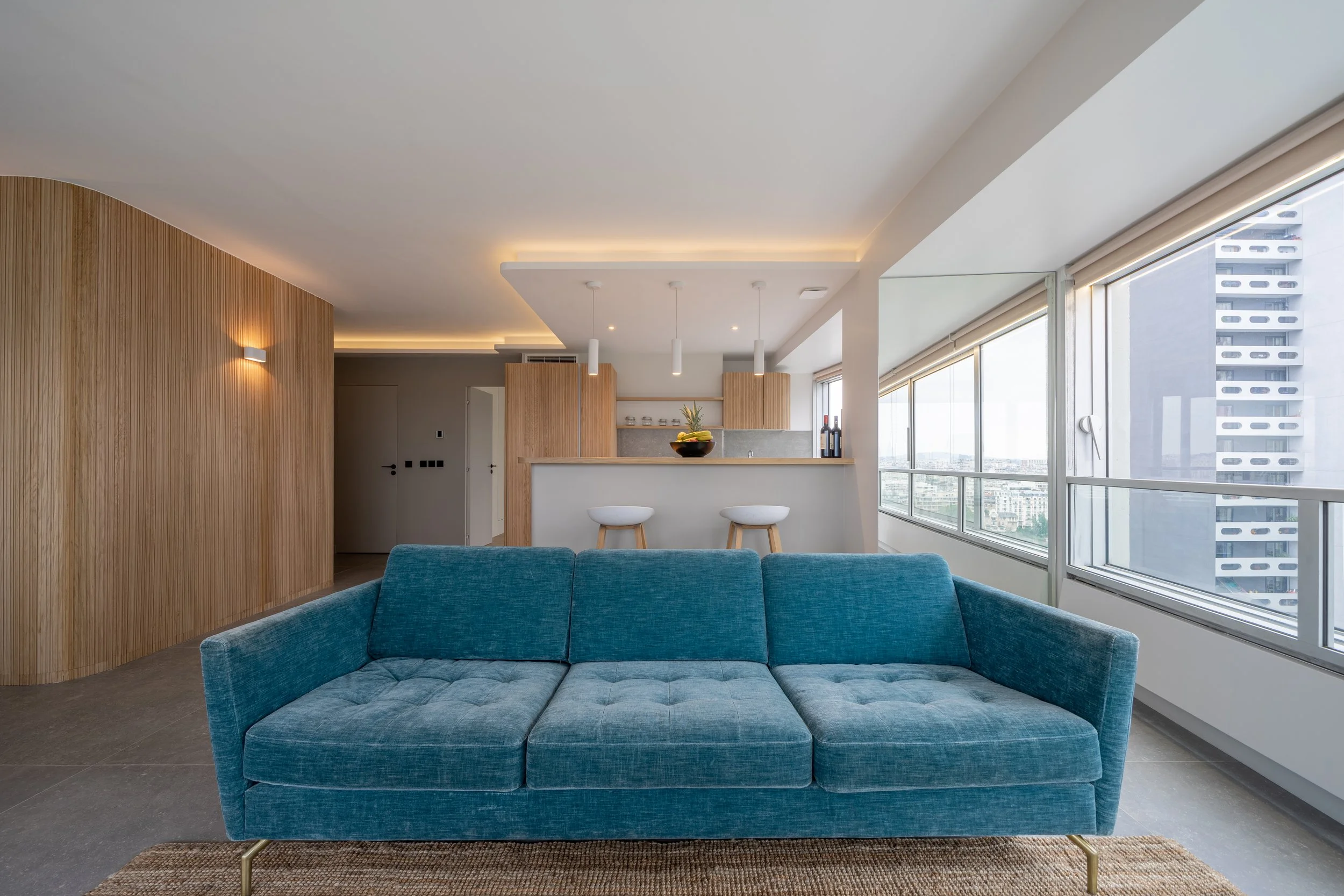 Interior of a modern apartment with a blue sofa, wooden wall paneling, and large windows showing a cityscape.