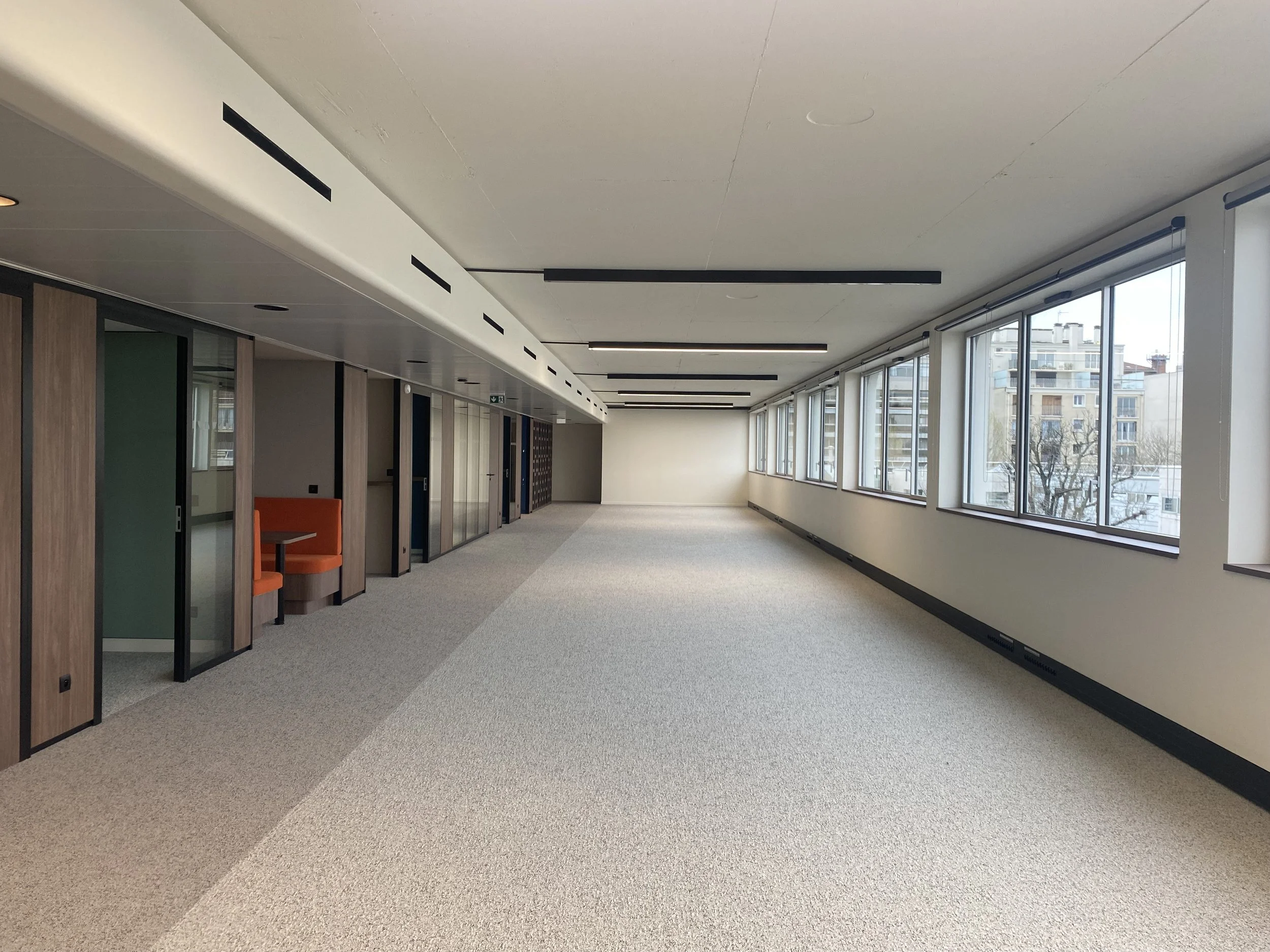 An empty modern hallway with large windows on the right side, allowing natural light and a cityscape view. On the left side, there are small private rooms or offices with glass doors framed in black and wood panels. The ceiling has long black linear light fixtures, and the floor is carpeted in neutral tones.