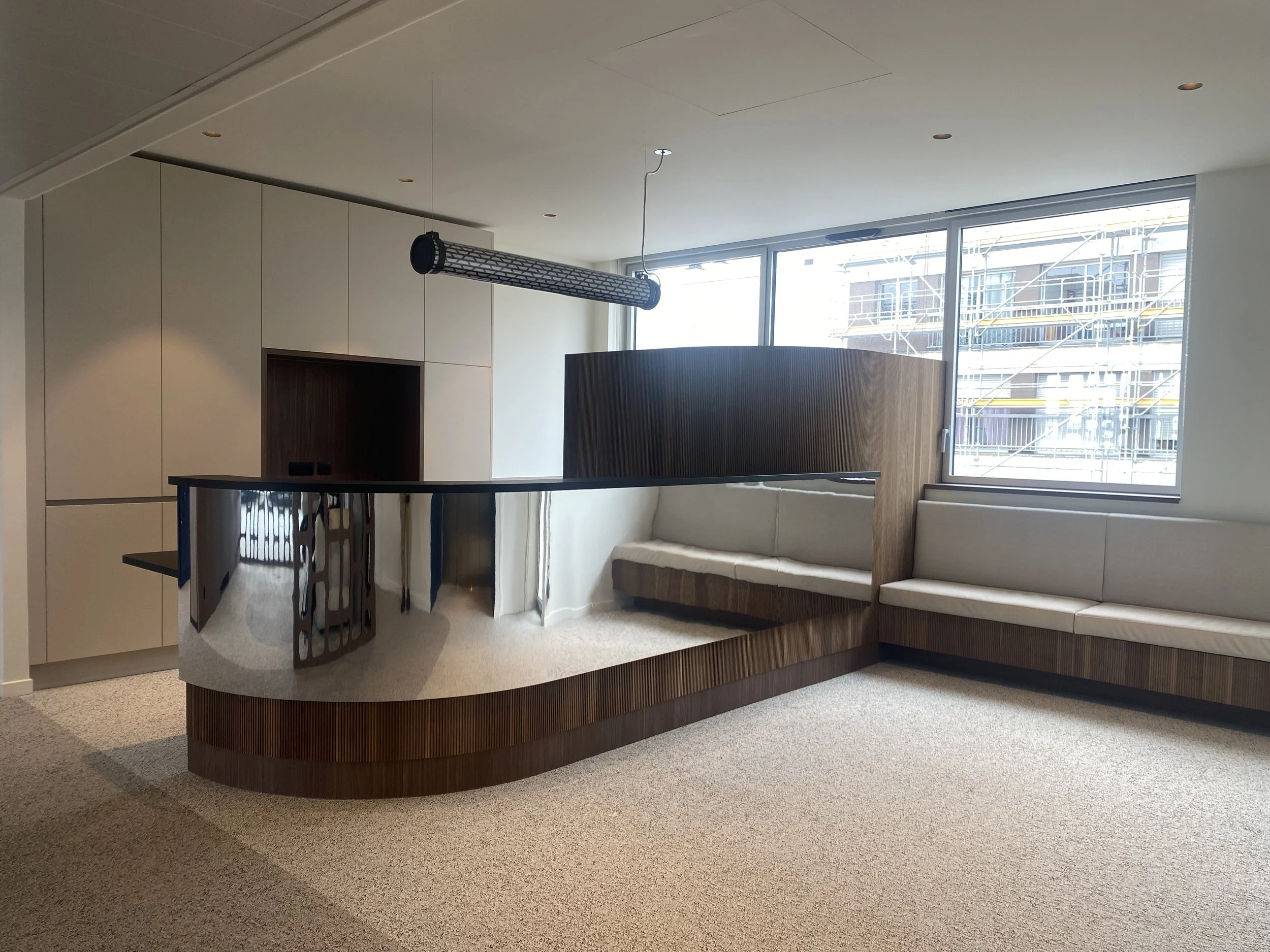 Interior of a modern room with a curved wooden bar, oil-rubbed dark wood and beige couches, large windows showing a construction site outside, and a hanging tube-shaped light fixture.