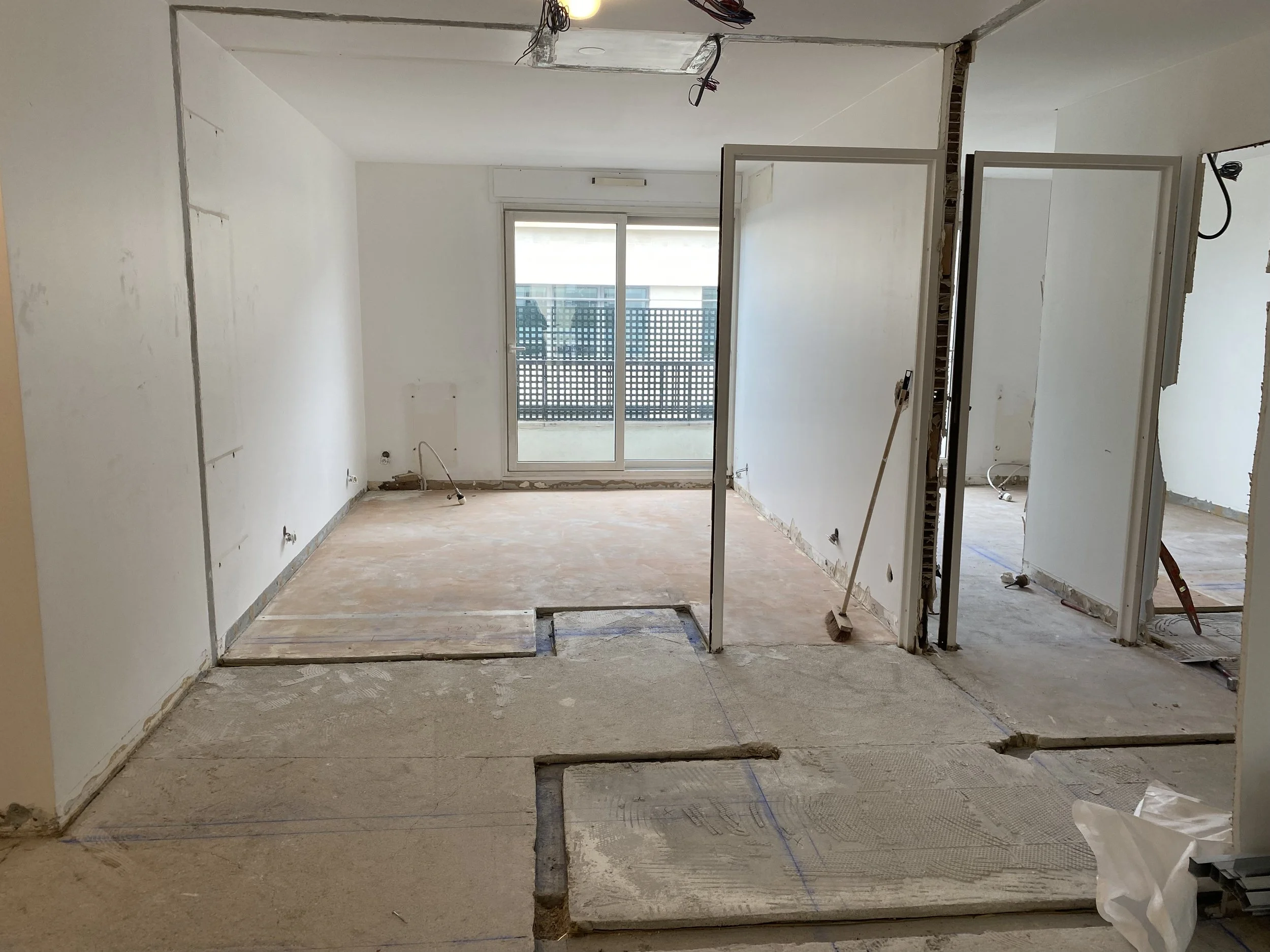 Room under renovation with unfinished flooring, exposed drywall, electrical wiring, and partially installed fixtures. A door leads outside with a balcony visible through the glass.