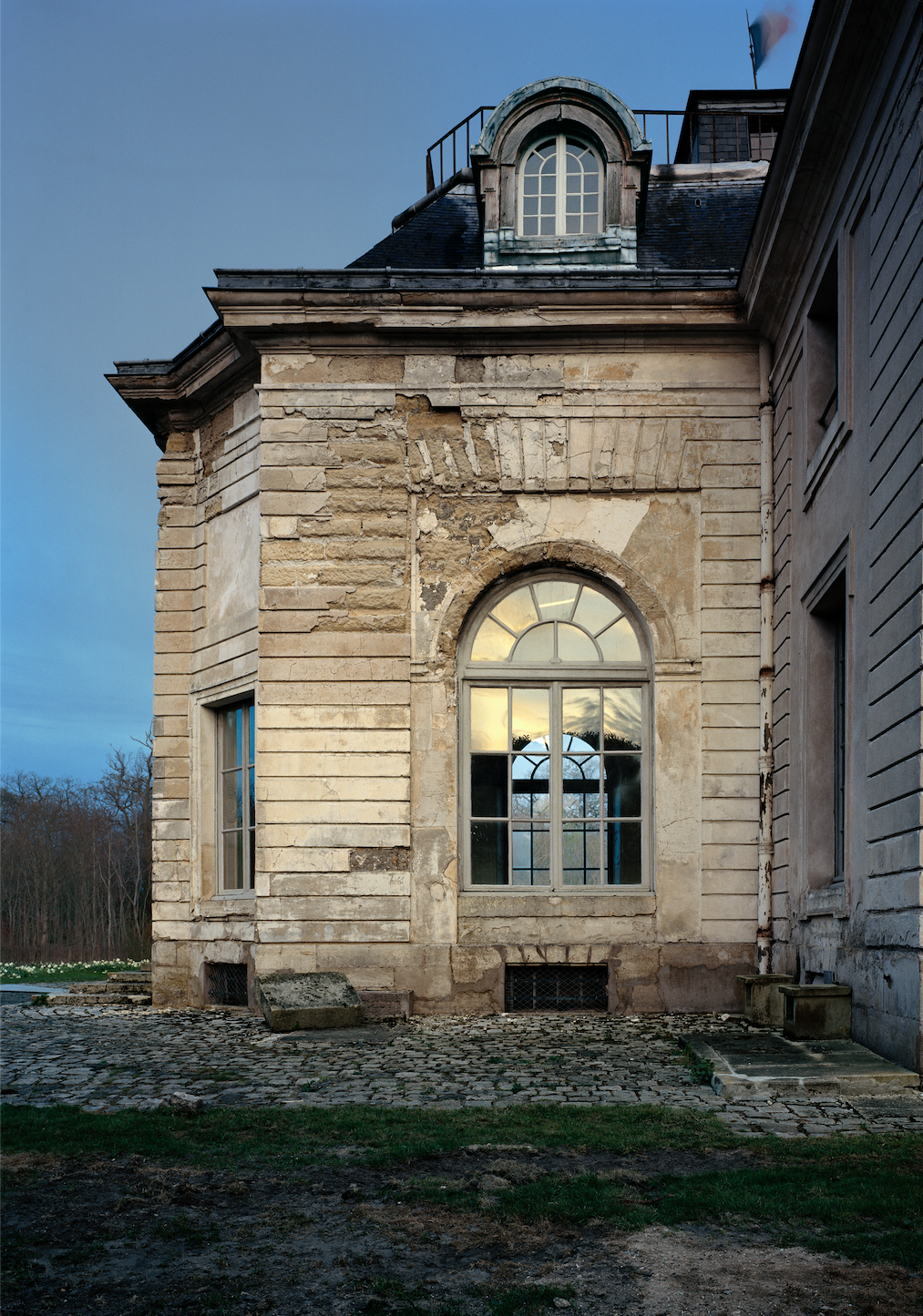 The side of an old, weathered stone building with arched windows, some with broken or missing panes, and a weathered roof. The building shows signs of neglect with peeling paint and damaged exterior walls.