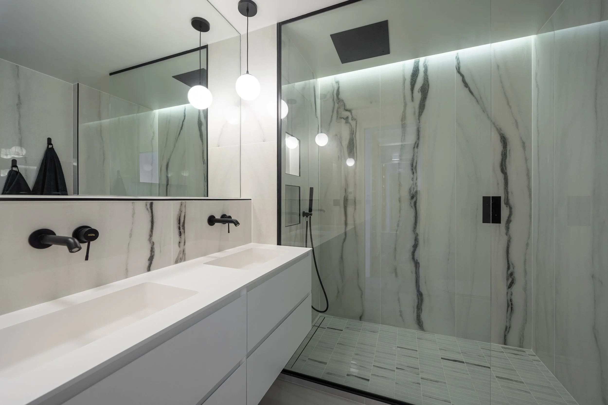 Modern bathroom with marble walls, a double white sink vanity, black fixtures, a large mirror, and a walk-in shower with a glass door and black fixtures.