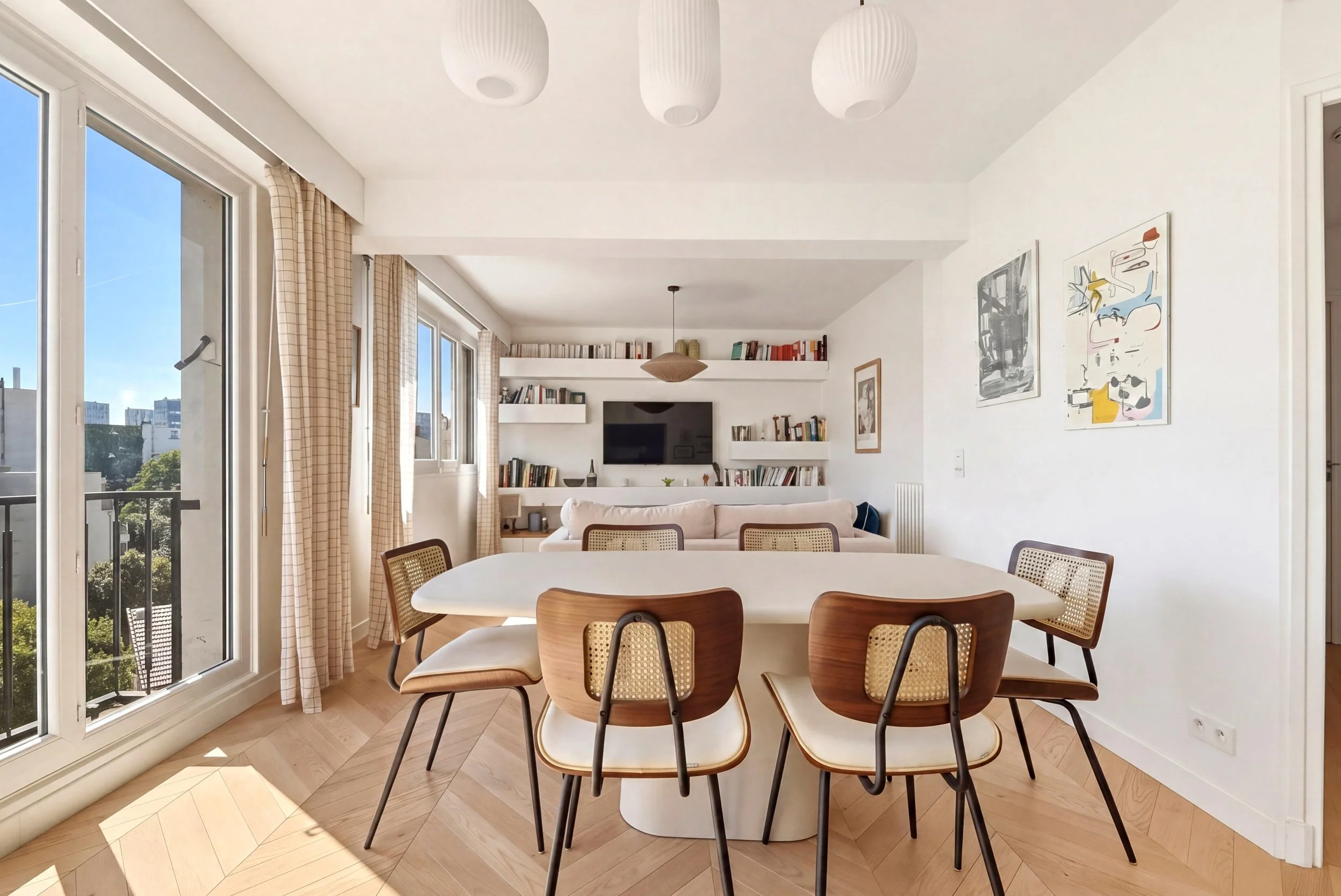 A bright and airy living and dining room with large windows and glass doors allowing in natural light. The room features a round dining table with wooden chairs and a cozy beige sofa in the background. White built-in shelves hold books and decorative items. Modern artwork decorates the walls, and white pendant lights hang from the ceiling. The space has a minimalist and contemporary design with light-colored hardwood flooring and beige curtains.