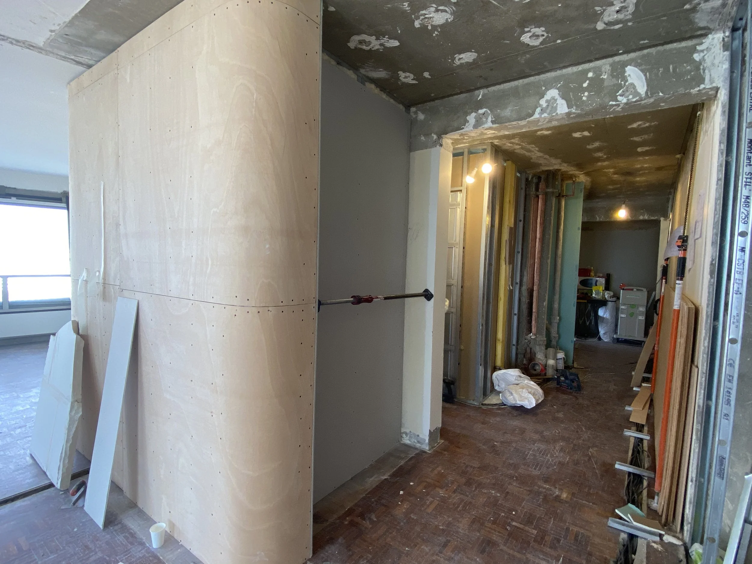 Interior of a building under construction, featuring a curved wooden wall panel, exposed ceiling with patches, scaffolding, construction tools and materials, and plastic pipes along the edge.