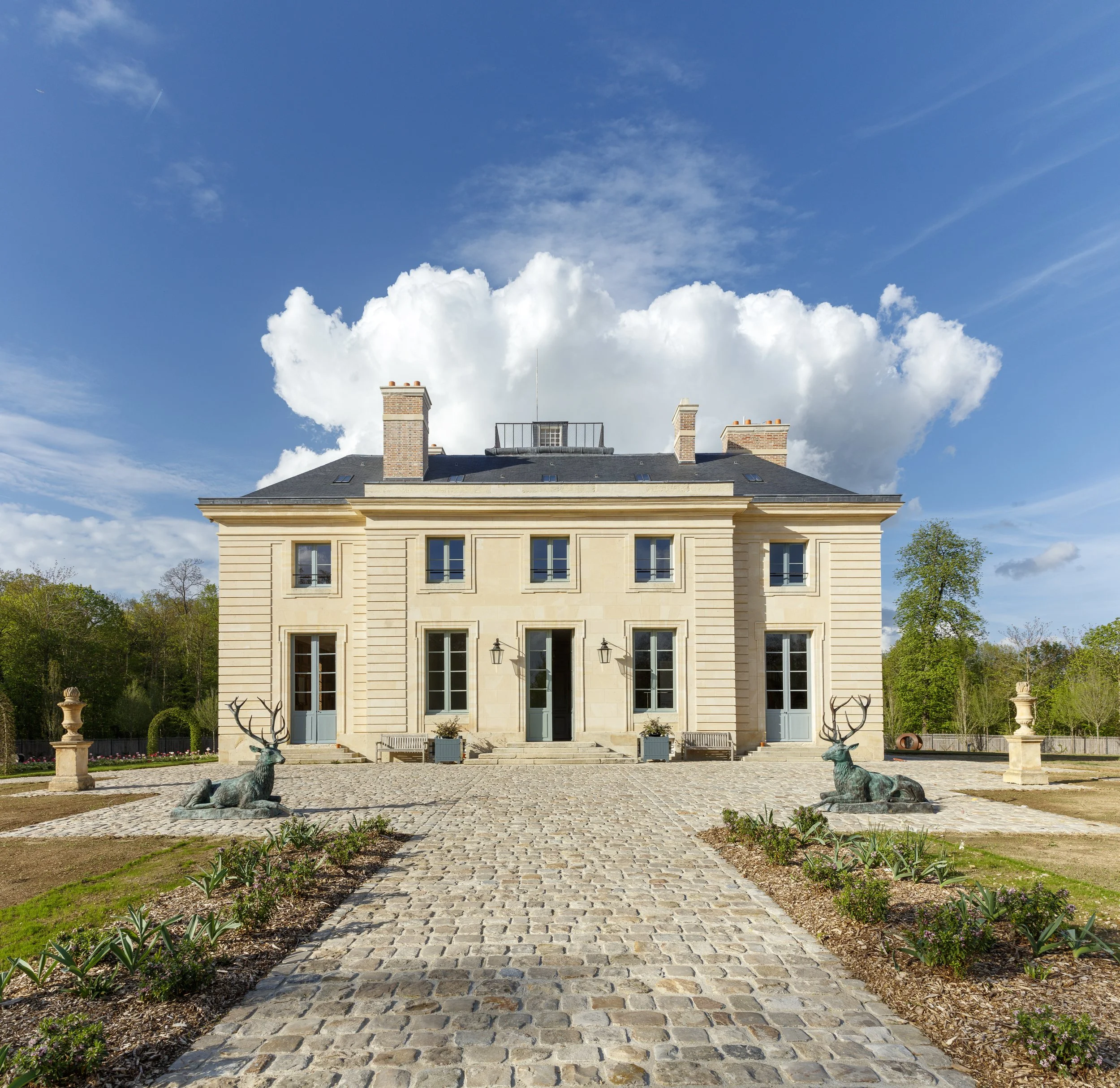 View of a stately house with a cobblestone pathway leading to the entrance, flanked by decorative sculptures of seated deer, green plants, and a lush garden landscape under a bright blue sky with scattered clouds.