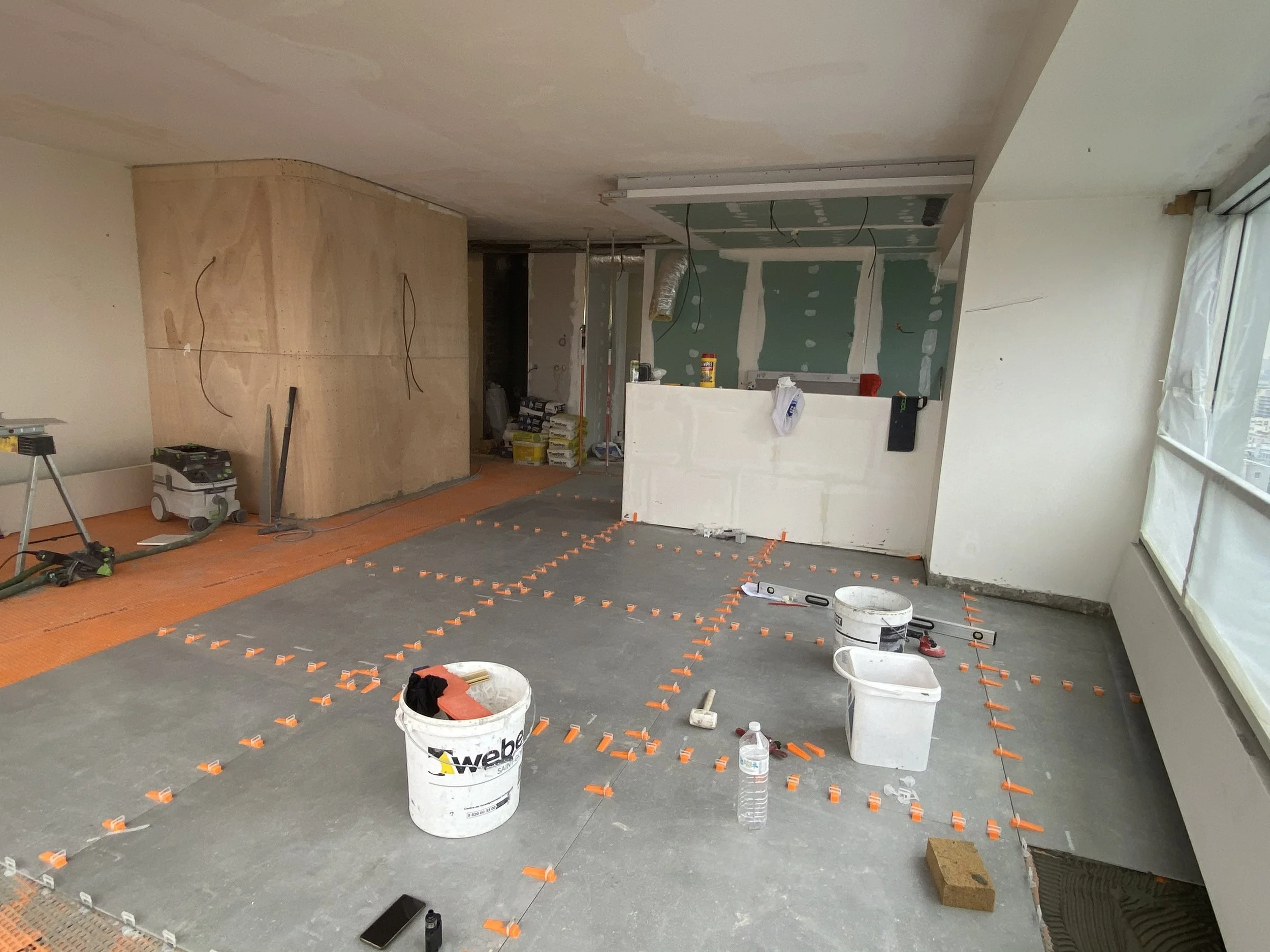 Room under construction with drywall and flooring being installed. Tools, buckets, and supplies are scattered around. Orange clips are used for securing flooring or underlayment.