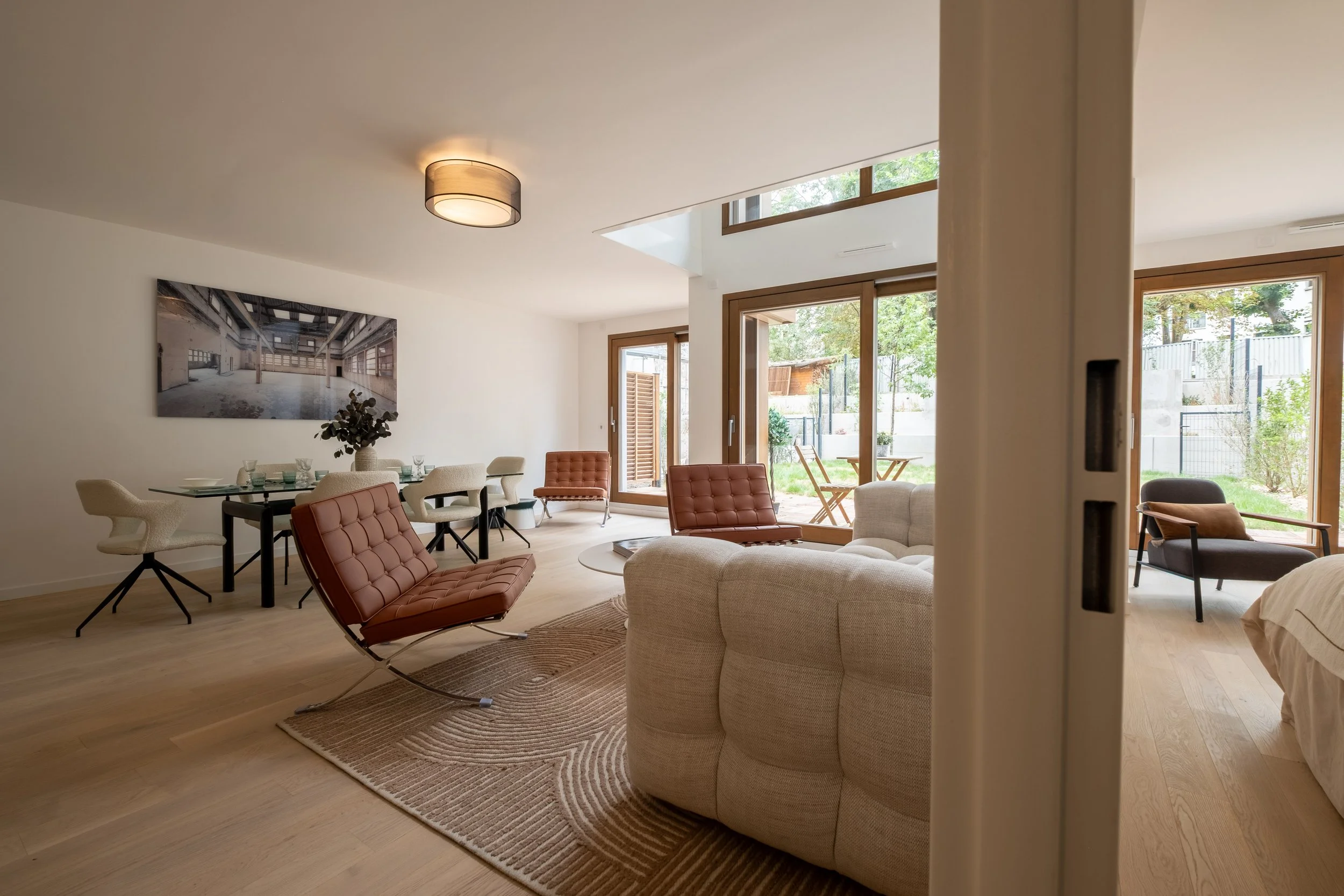 Modern living room with beige sofa, brown leather chairs, a round wooden coffee table, a dining table with beige chairs, large glass doors and windows, and outdoor greenery visible.