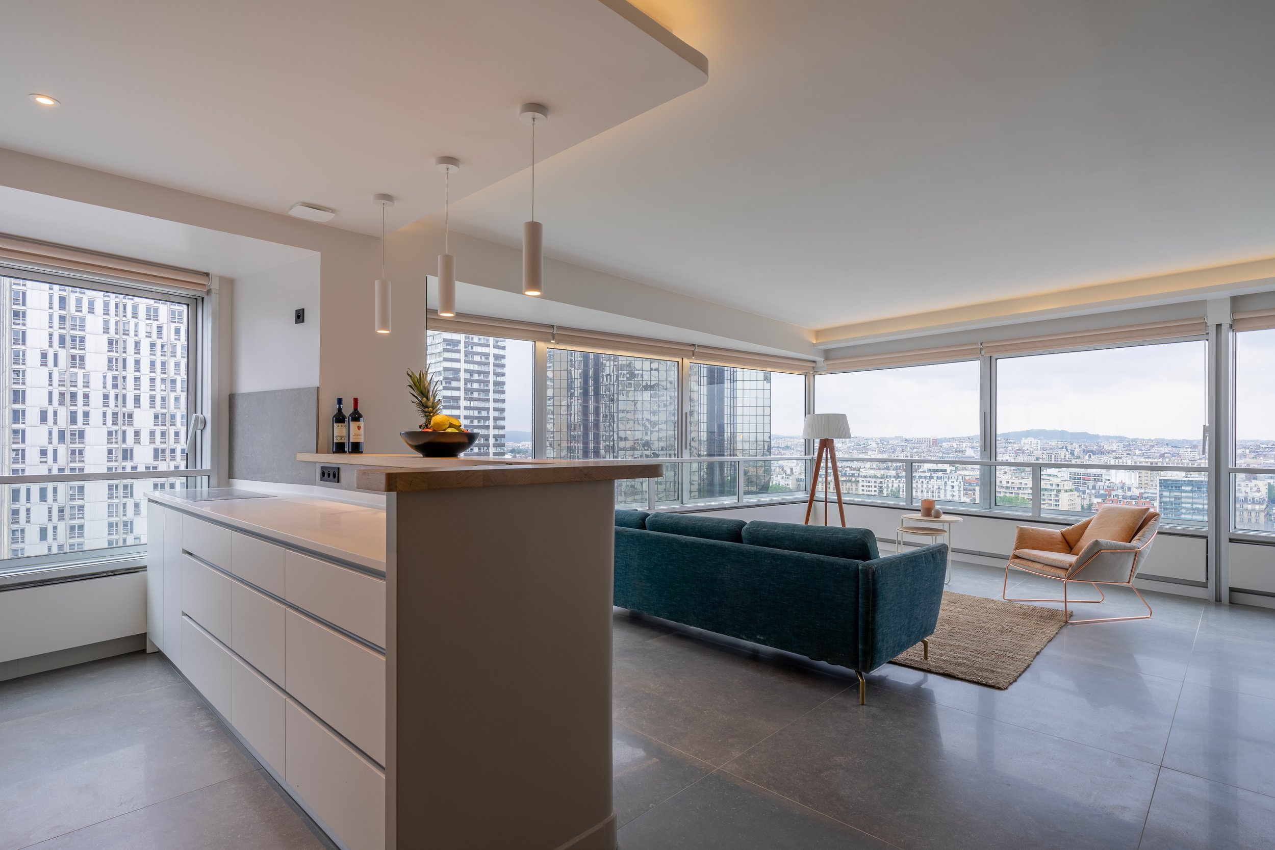 Modern high-rise apartment living room with large windows showing city view, featuring a navy blue sofa, a beige armchair, a standing lamp, and a small side table with decor.