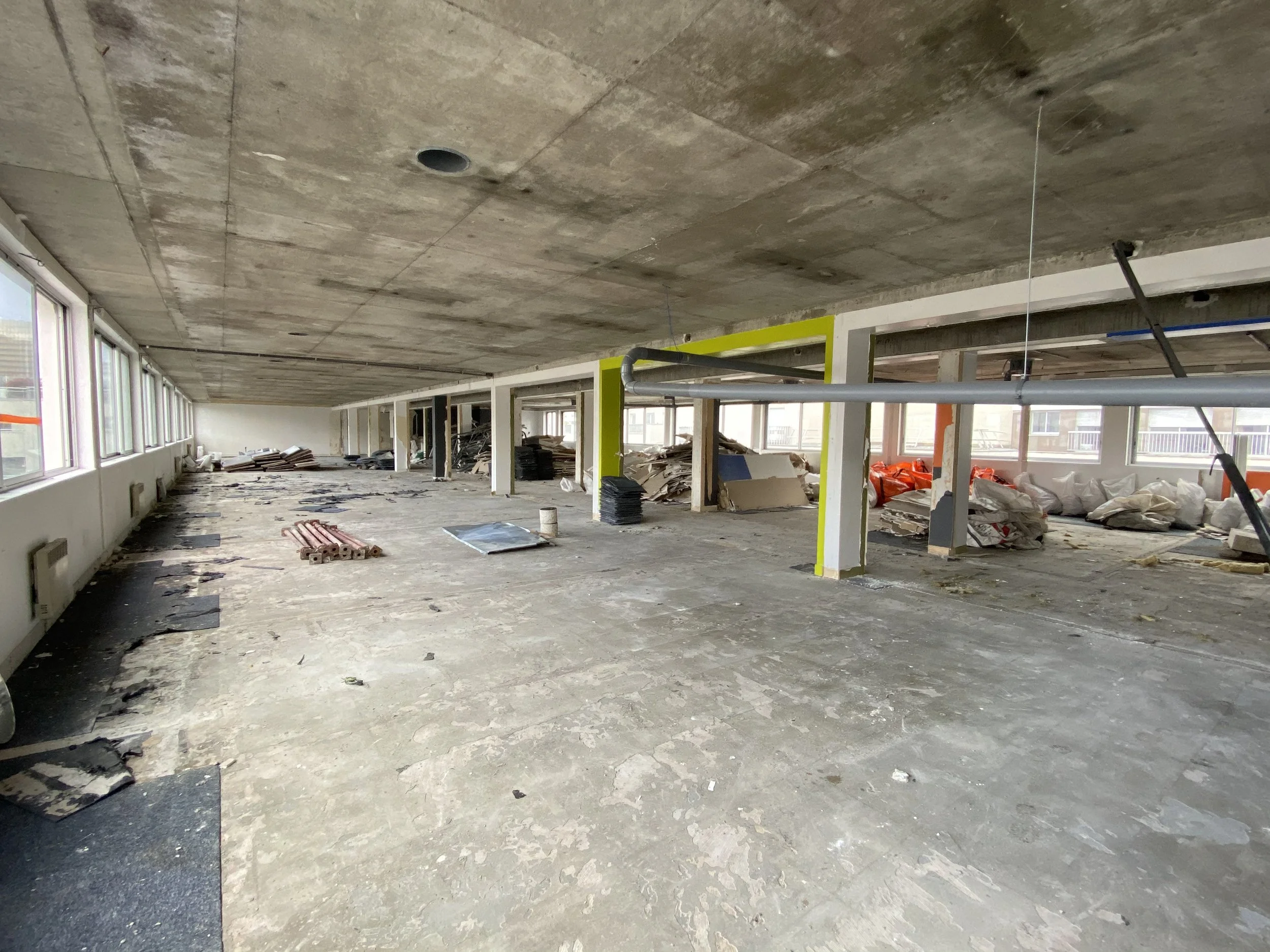 Interior of a building under construction, with exposed concrete ceiling and floor, construction materials, and debris scattered around. Multiple windows line one wall, allowing natural light into the space.