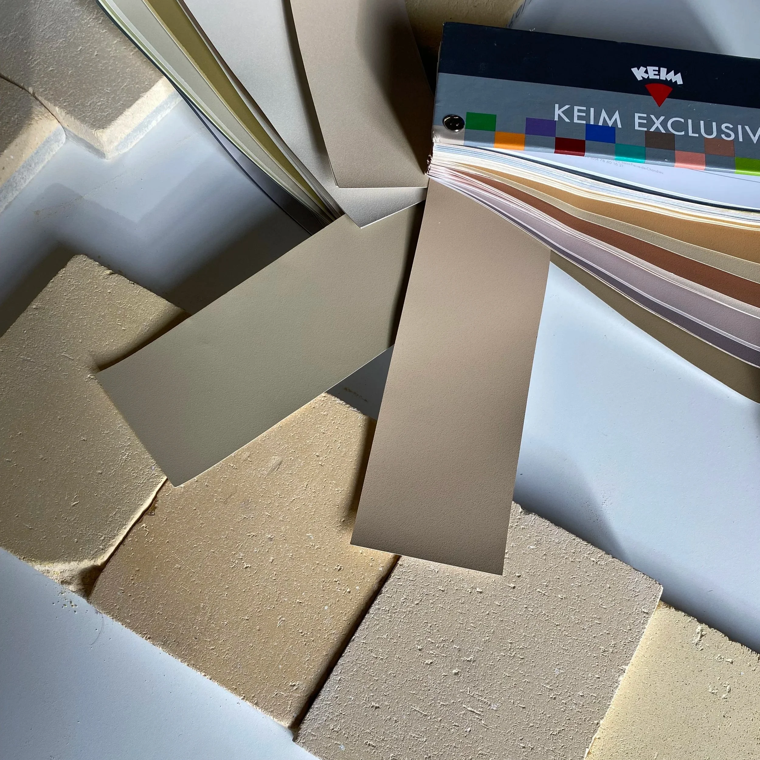 Samples of various textured and colored wall materials, including paint and plaster, arranged in overlapping sheets with a KEM EXCLUSIV branding booklet placed among them.