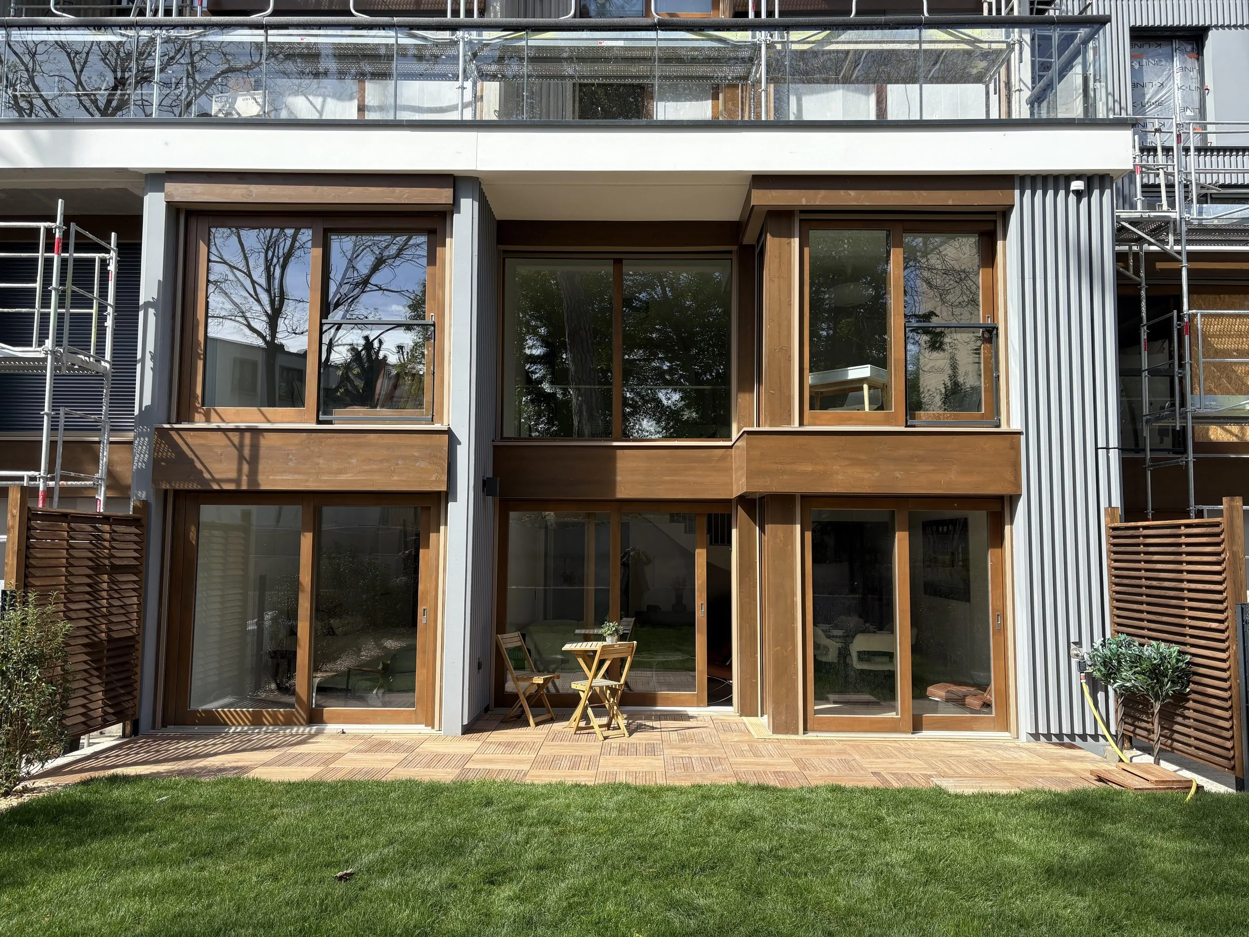 Modern multi-story building under construction with large glass windows and wooden accents, scaffolding on the sides, and a small patio area with a table and chair in the backyard.
