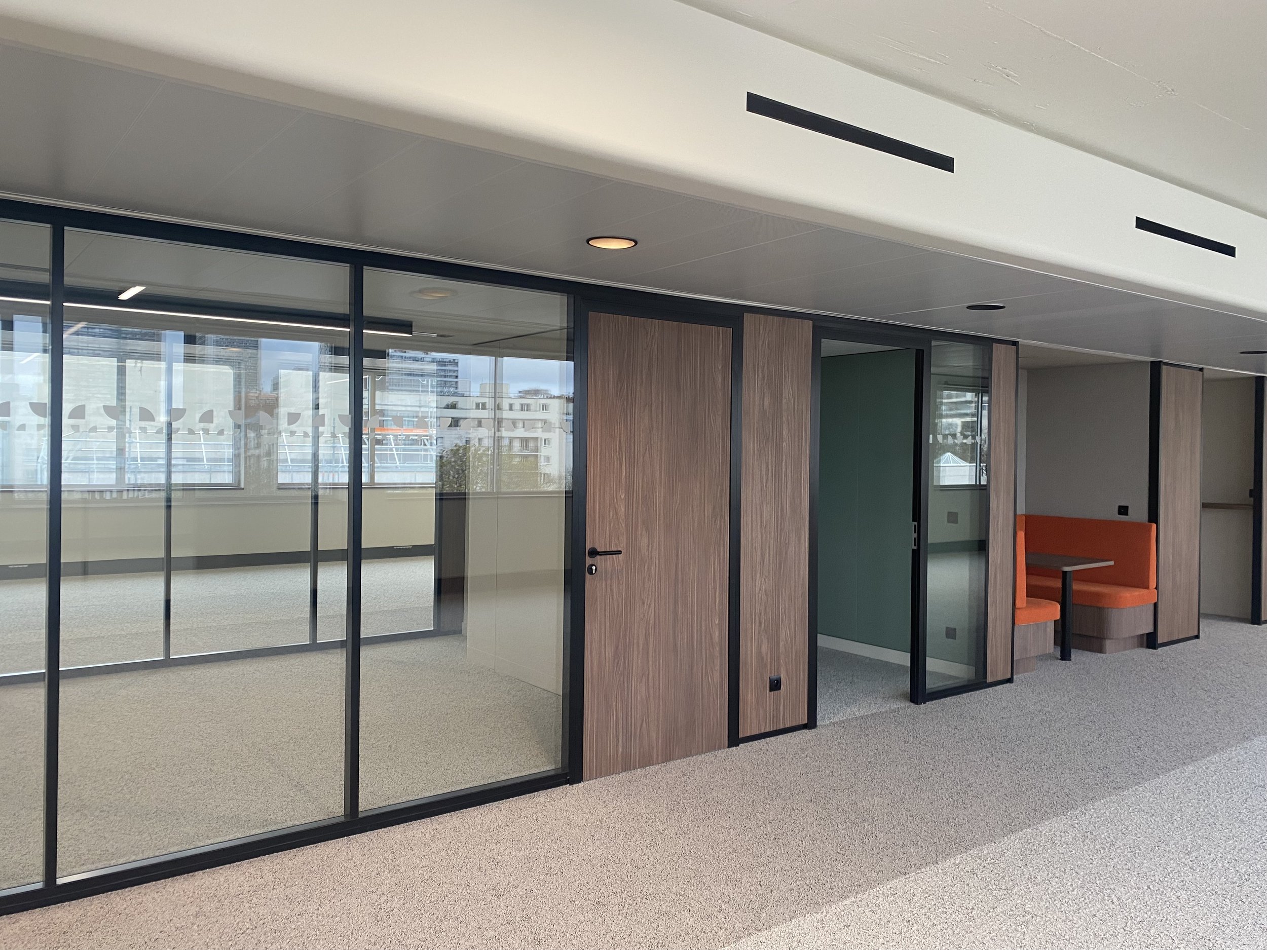 Interior view of a modern office space with glass partitions, wooden doors, and orange seating. Large windows in the background reveal city buildings.