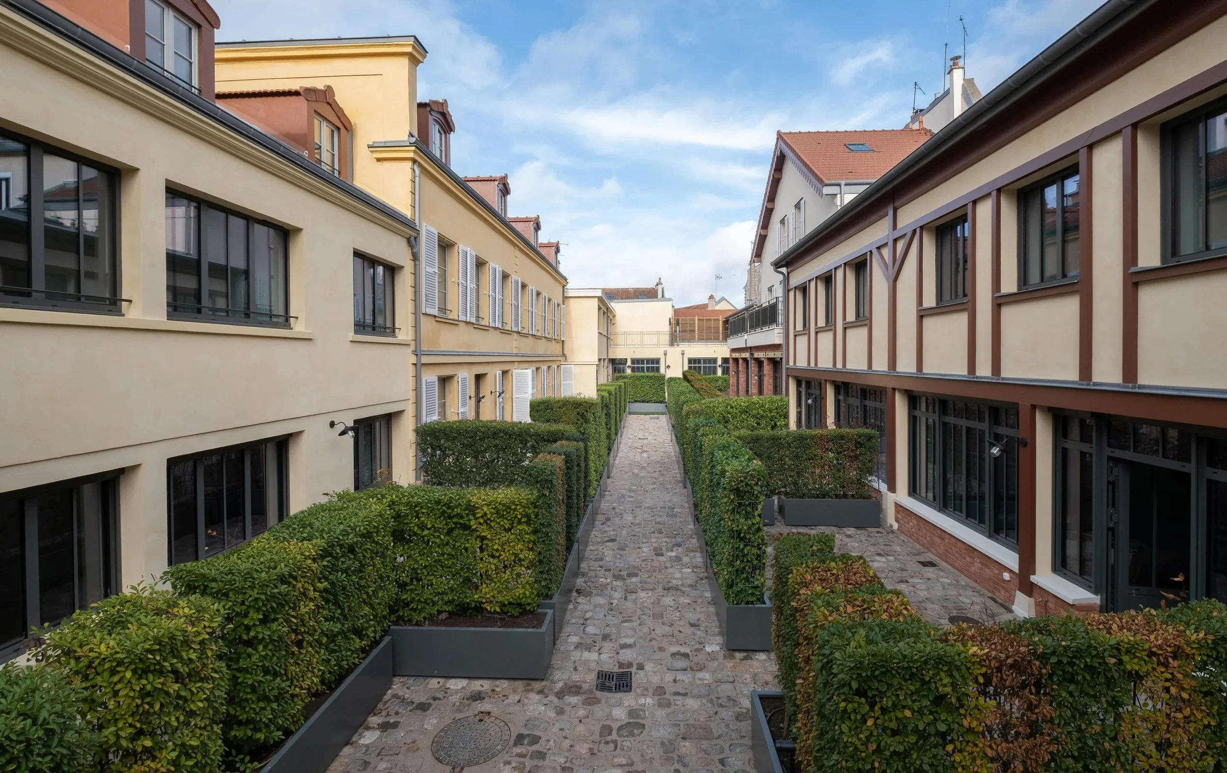 View of a narrow cobblestone walkway between modern apartment buildings with beige and brown facades, green bushes in planters, and a blue sky with some clouds.