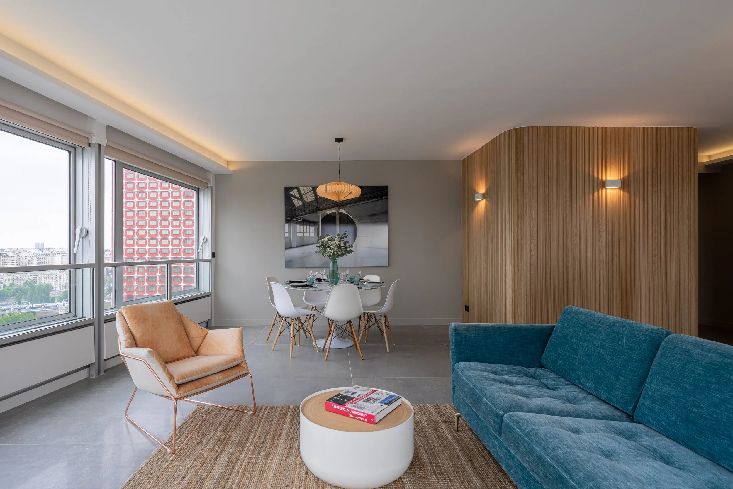 Modern living room with large windows, a blue sofa, a peach chair, a white round coffee table with books, a dining table with white chairs, wall lighting, and a wooden accent wall.