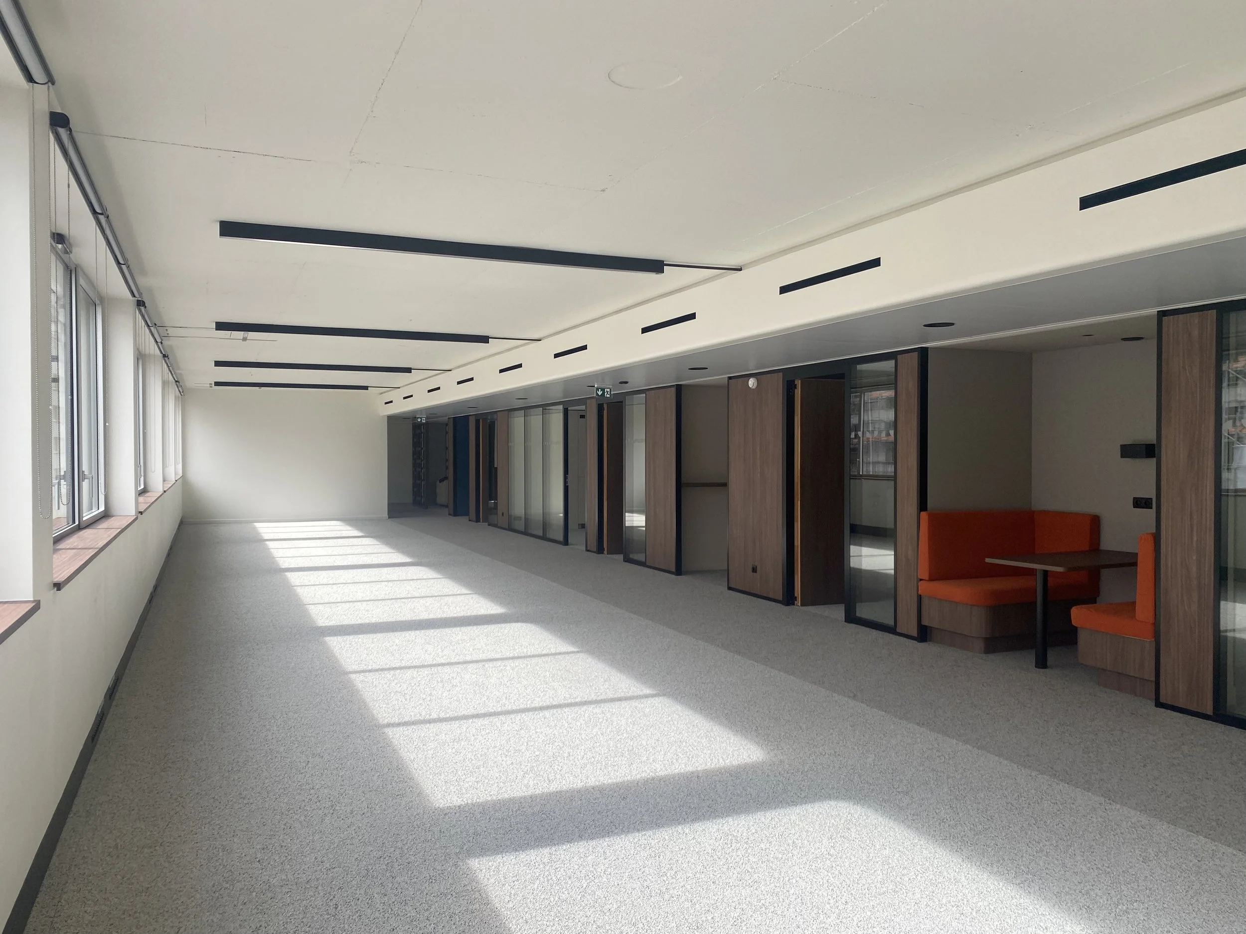An empty modern office corridor with large windows on the left, white walls, and a ceiling with black linear light fixtures. On the right, there are glass-walled offices with wooden doors and an orange sofa.