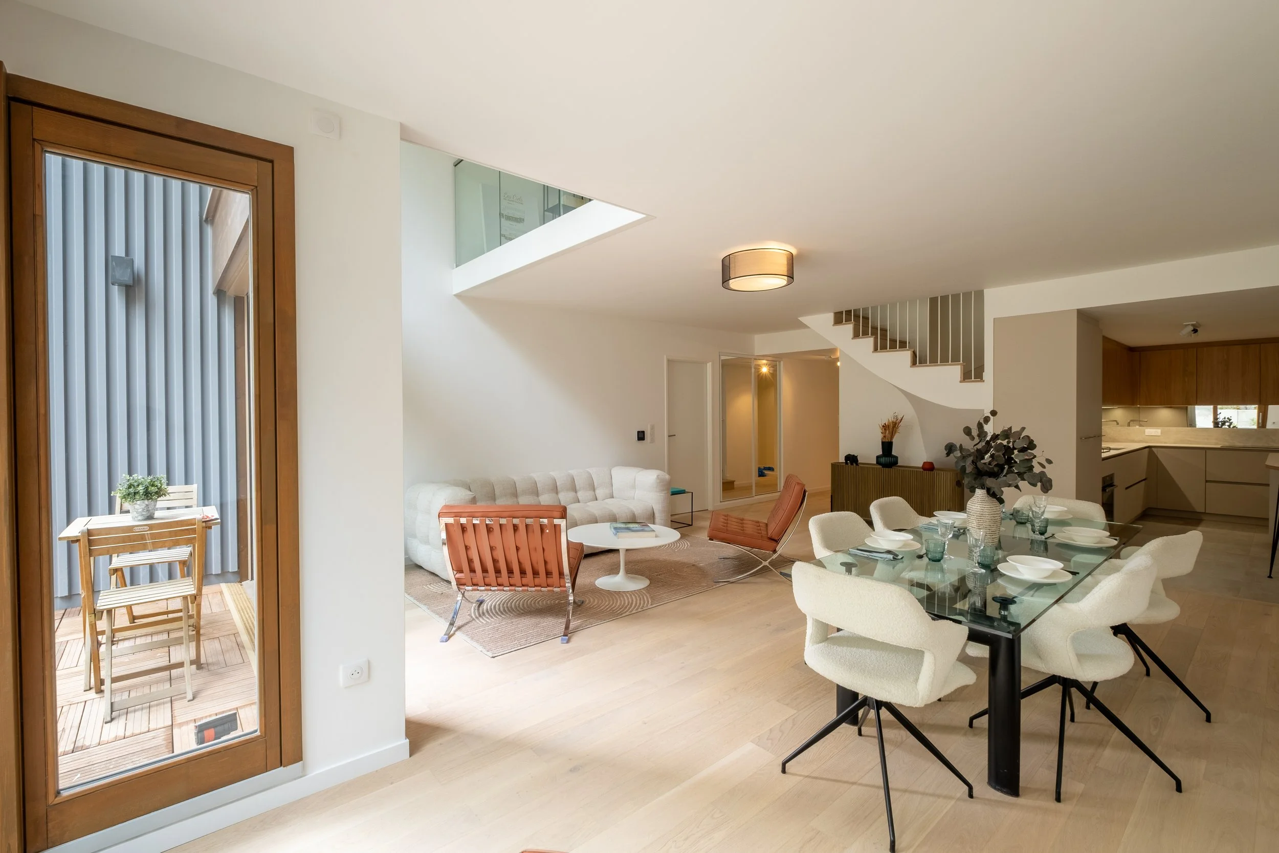 Modern open-concept living and dining area with a glass dining table, white upholstered chairs, beige sofa, mid-century modern chairs, and sliding glass door leading to a small balcony with wooden flooring and outdoor furniture.